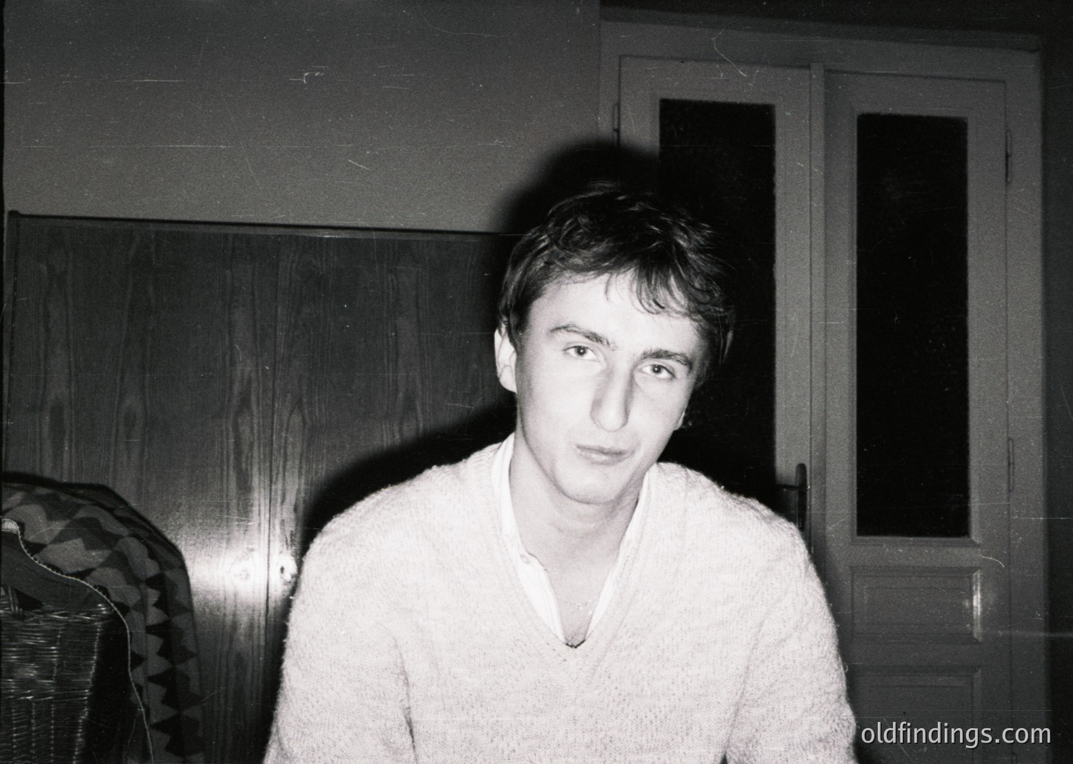 Portrait of a young man in a light sweater, captured in low-light indoor setting. Wooden door and patterned upholstery suggest mid-20th century domestic interior. Expression neutral, composition emphasizes candid realism.