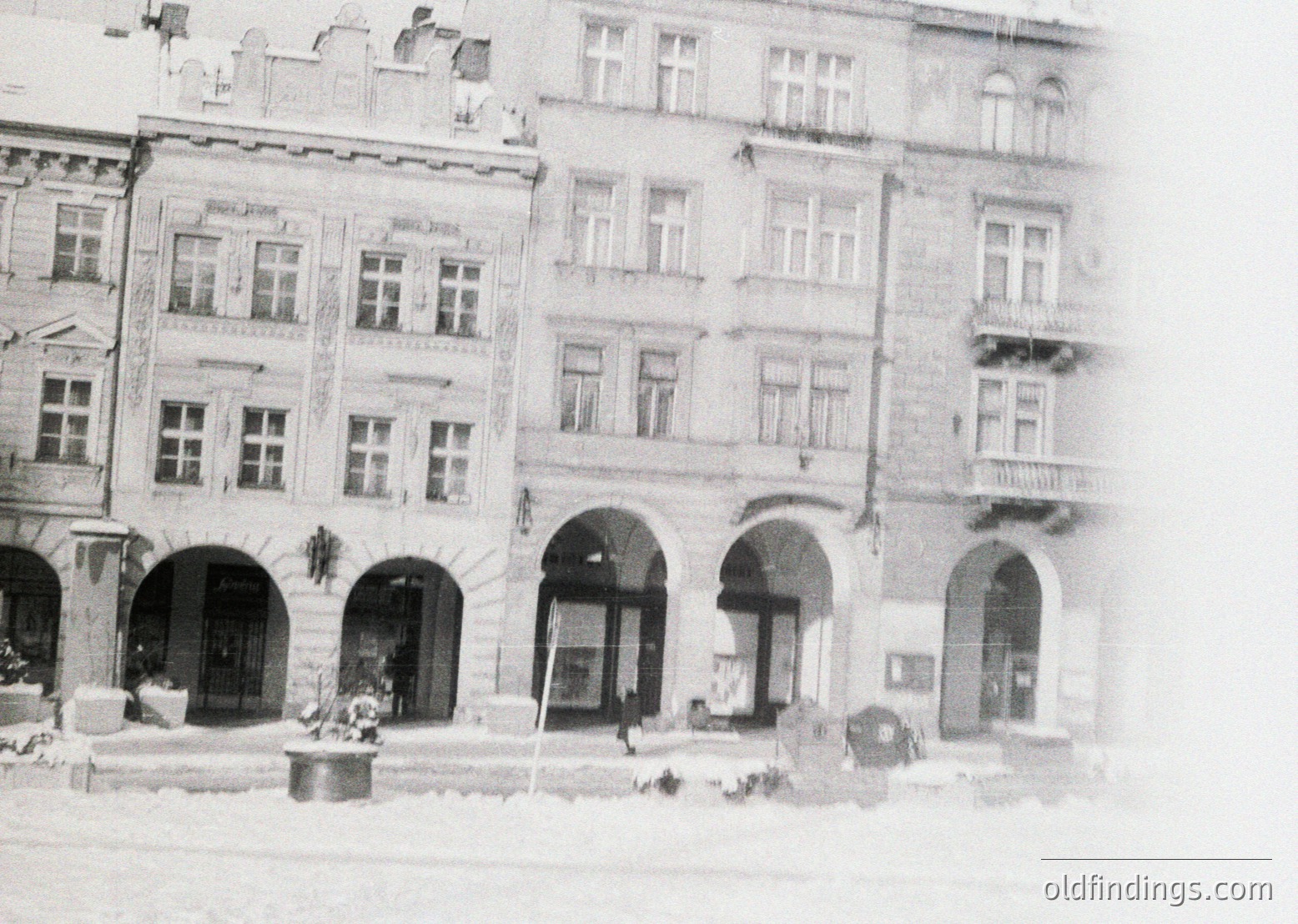 Blurred vintage street scene featuring three-story European-style buildings with arched entrances, decorative cornices, and large rectangular windows. Snow covers rooftops and ground, indicating winter. Signage suggests commercial use ( )