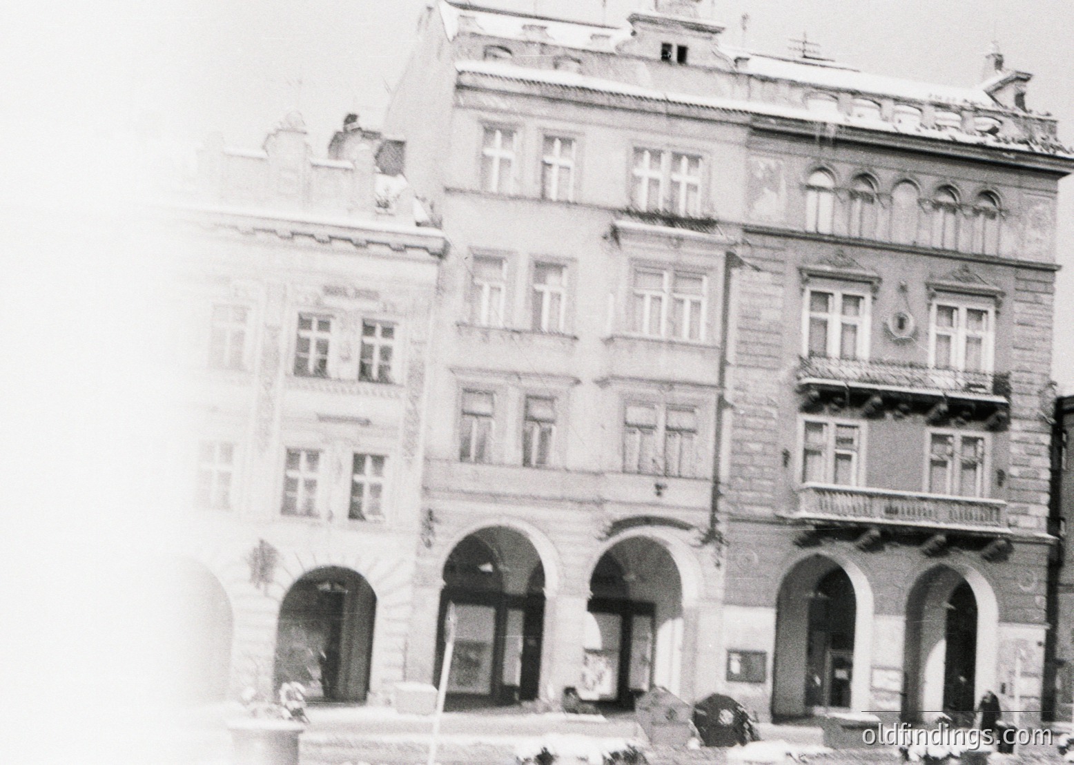 Three-story European building with Art Nouveau/Beaux-Arts details—arched ground-floor entrances, decorative stonework, and wrought-iron balconies. Likely late 19th to early 20th century. Urban street scene with pedestrians and vintage vehicles.