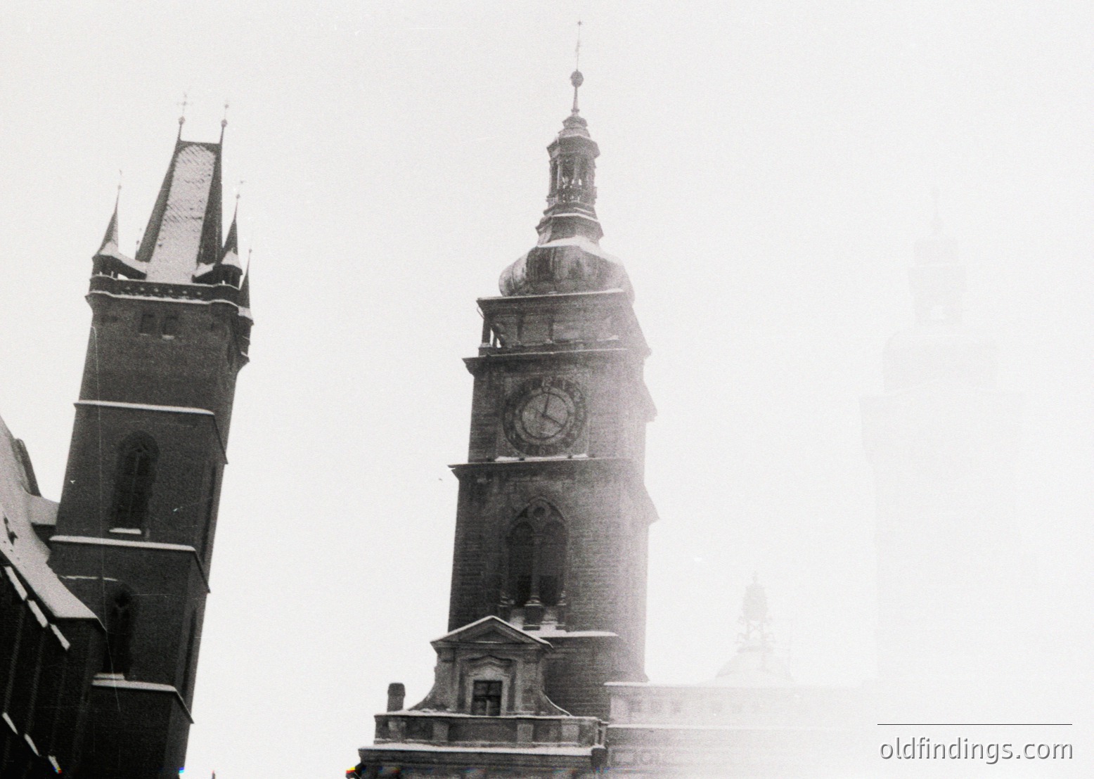 Black-and-white photograph of a historic European clock tower with Gothic Revival architecture, featuring a prominent round clock face and ornate spire. Adjacent church steeple with pointed roof in misty background. Likely 19th-century urban setting.