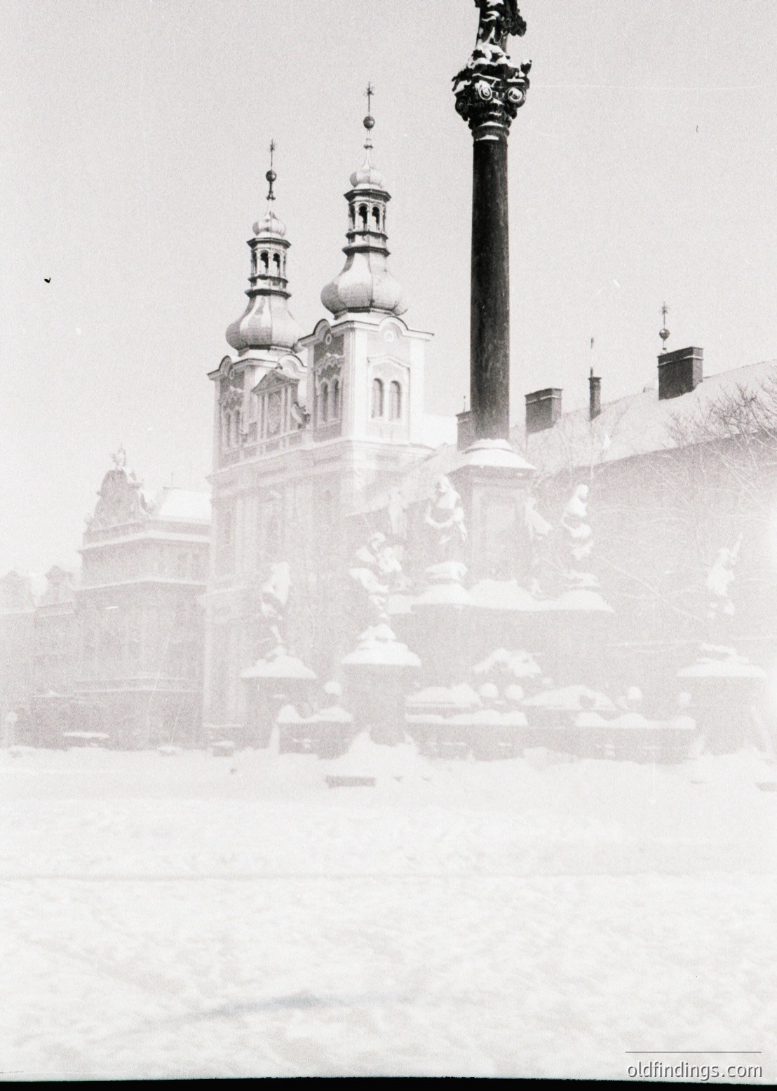 Historic Baroque-style cathedral with twin domed towers partially obscured by mist/snow. Ornate façade and decorative column in foreground. Likely European winter setting, mid-20th century.