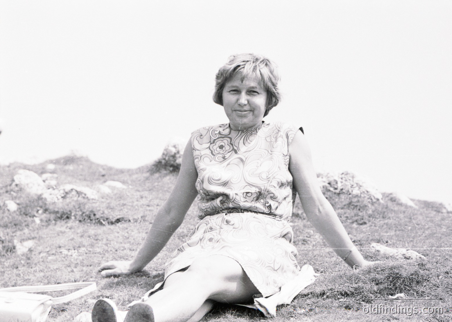 Portrait of a woman in a floral-patterned dress, seated outdoors on grassy terrain with rocky background. Mid-20th century fashion, likely 1960s.