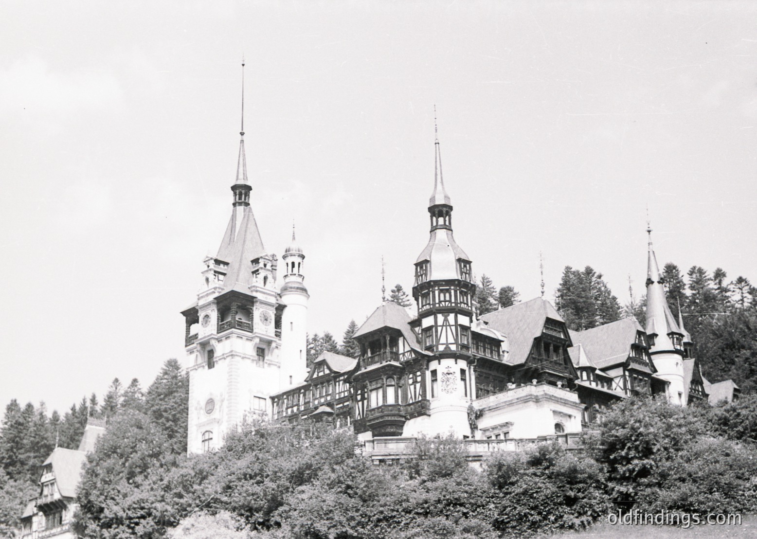 Historic castle with Gothic Revival architecture, featuring steeply pitched roofs, turrets, and ornate detailing. Surrounded by dense forest, likely Romania’s **Peleș Castle**, built 1873–1883.