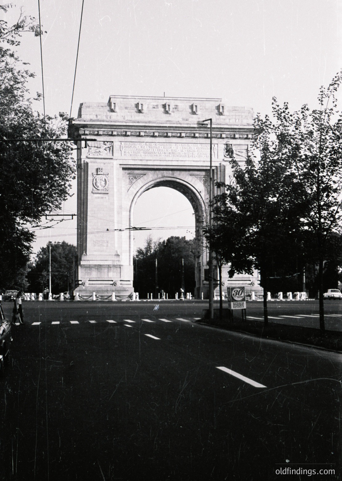 Neoclassical triumphal arch with intricate carvings and decorative columns, flanked by trees and street lamps. Likely a historical landmark in an urban setting, possibly ’s Puerta de Alcalá (18th century). Symmetrical design and grand scale evoke imperial architecture. á