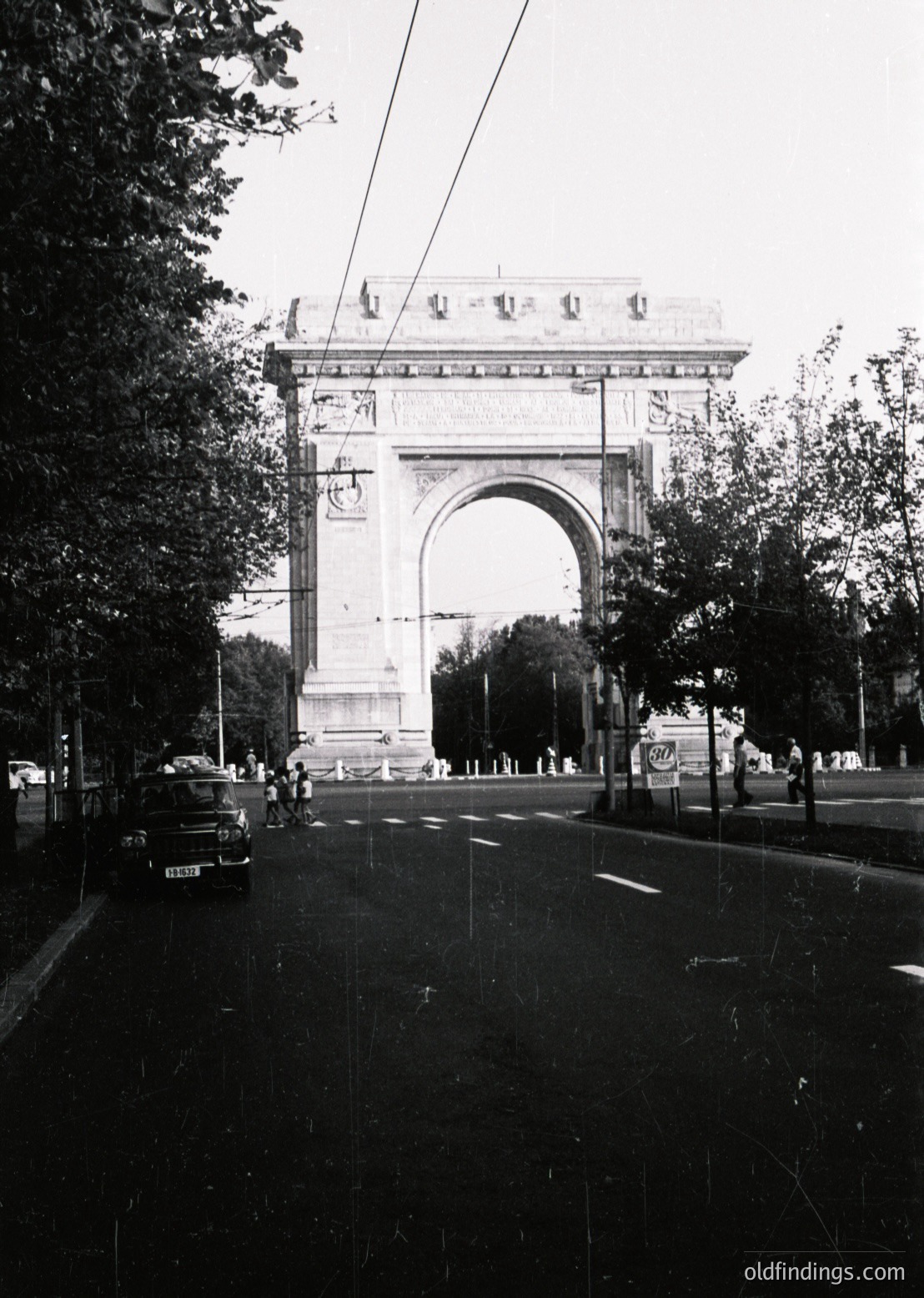 Neoclassical triumphal arch with intricate carvings and a central inscription, flanked by statues. Urban street scene with vintage car and pedestrians, suggesting mid-20th century. Likely a European city center.