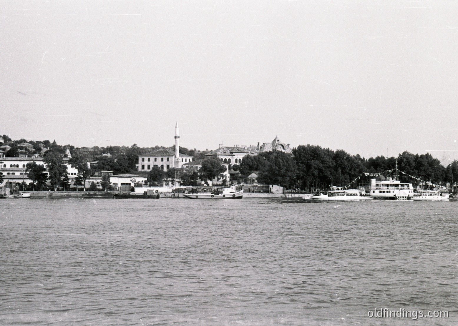 Black-and-white coastal scene featuring a prominent **minaret** and **waterfront buildings** along a shoreline. Small **ferries** and boats docked near a **wooden pier**. Dense **trees** and **low-rise architecture** suggest a historic port town. Likely **Istanbul, Turkey**, mid-20th century.