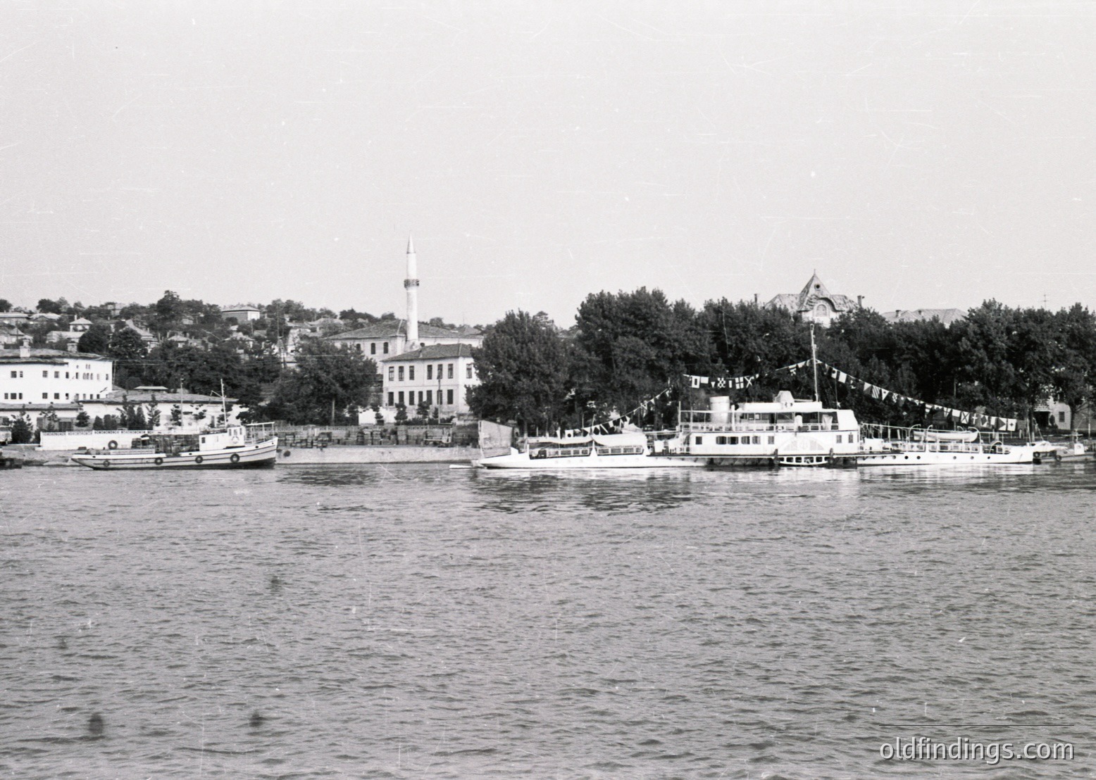 Black-and-white coastal scene featuring a waterfront promenade with a prominent building featuring a tall minaret and flagpole. Small boats navigate the harbor, while festive flags line the shore. Likely Istanbul, 1950s–1960s.