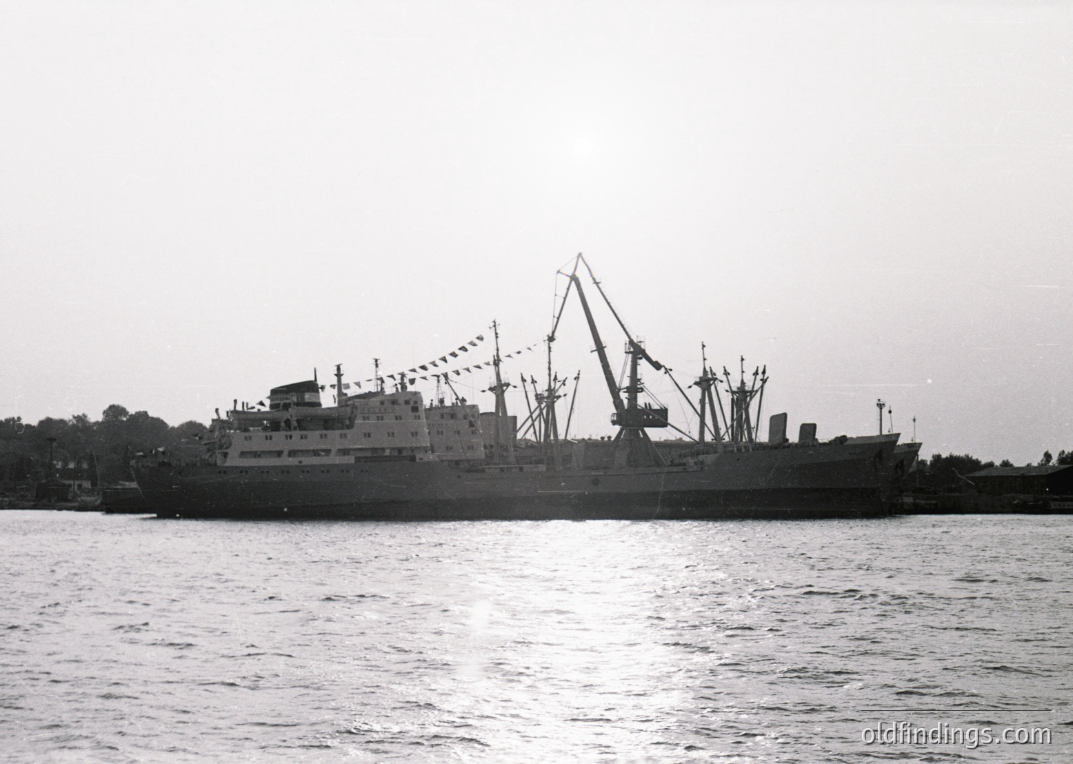 Mid-20th century cargo ship with cranes, likely a Soviet-era bulk carrier or ferry, sailing on calm waters. Decorative flags along the railing suggest a special occasion or voyage. Industrial port or coastal setting.