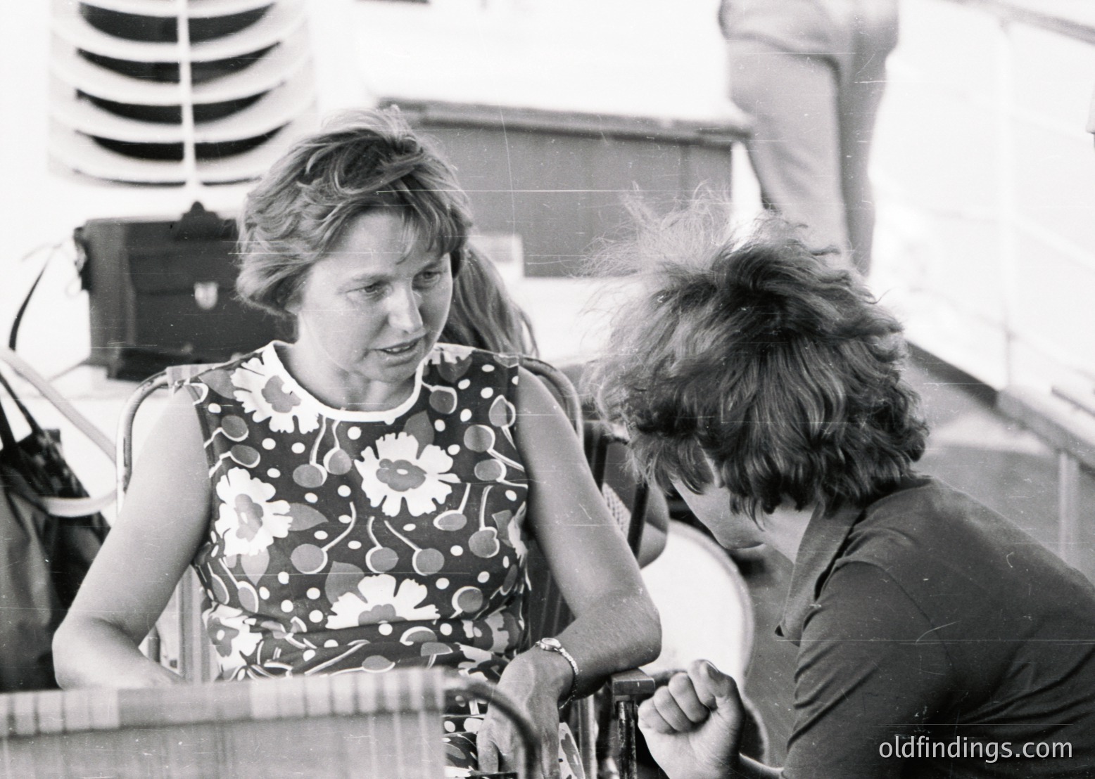 Black-and-white candid shot of two individuals engaged in conversation on a boat deck, likely mid-20th century. Woman in floral-patterned sleeveless dress with short hair; man in dark polo shirt leans in, gesturing. Industrial equipment (speaker, reel-to-reel tape recorder) in background suggests live audio recording. Casual, informal setting—possibly a maritime event or festival.