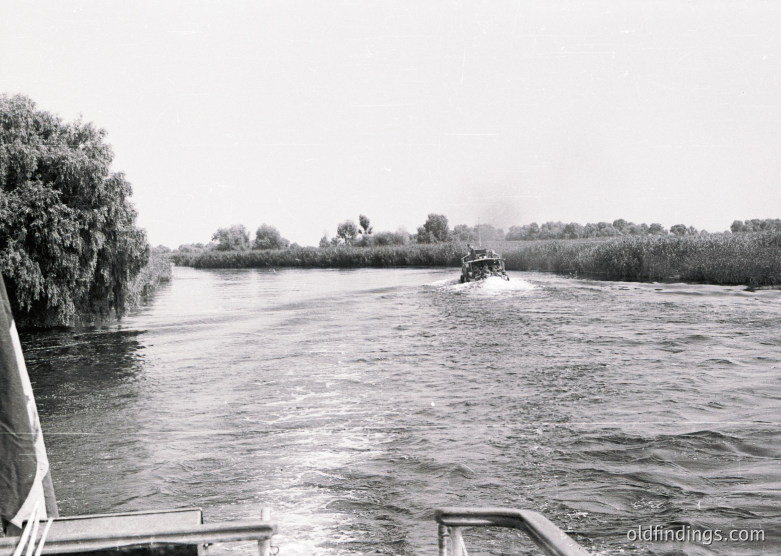 Vintage black-and-white photo of a small motorboat navigating a narrow, reed-lined waterway. Overcast skies and rippled water suggest mid-20th century (1950s–1970s) European river transport. Boat’s open cabin and simple design reflect utilitarian river travel.