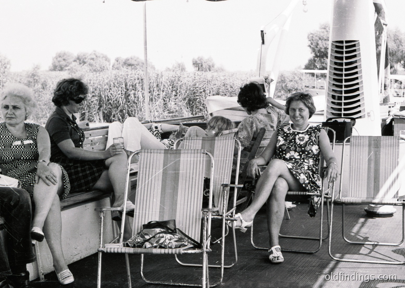 Black-and-white photo of four women relaxing on vintage deck chairs on a sun deck, likely mid-20th century. Two women in floral dresses and one in a checkered blouse sit, while a child reclines on a chair. Background shows greenery and a tall vertical structure, possibly a lifeguard tower or antenna.