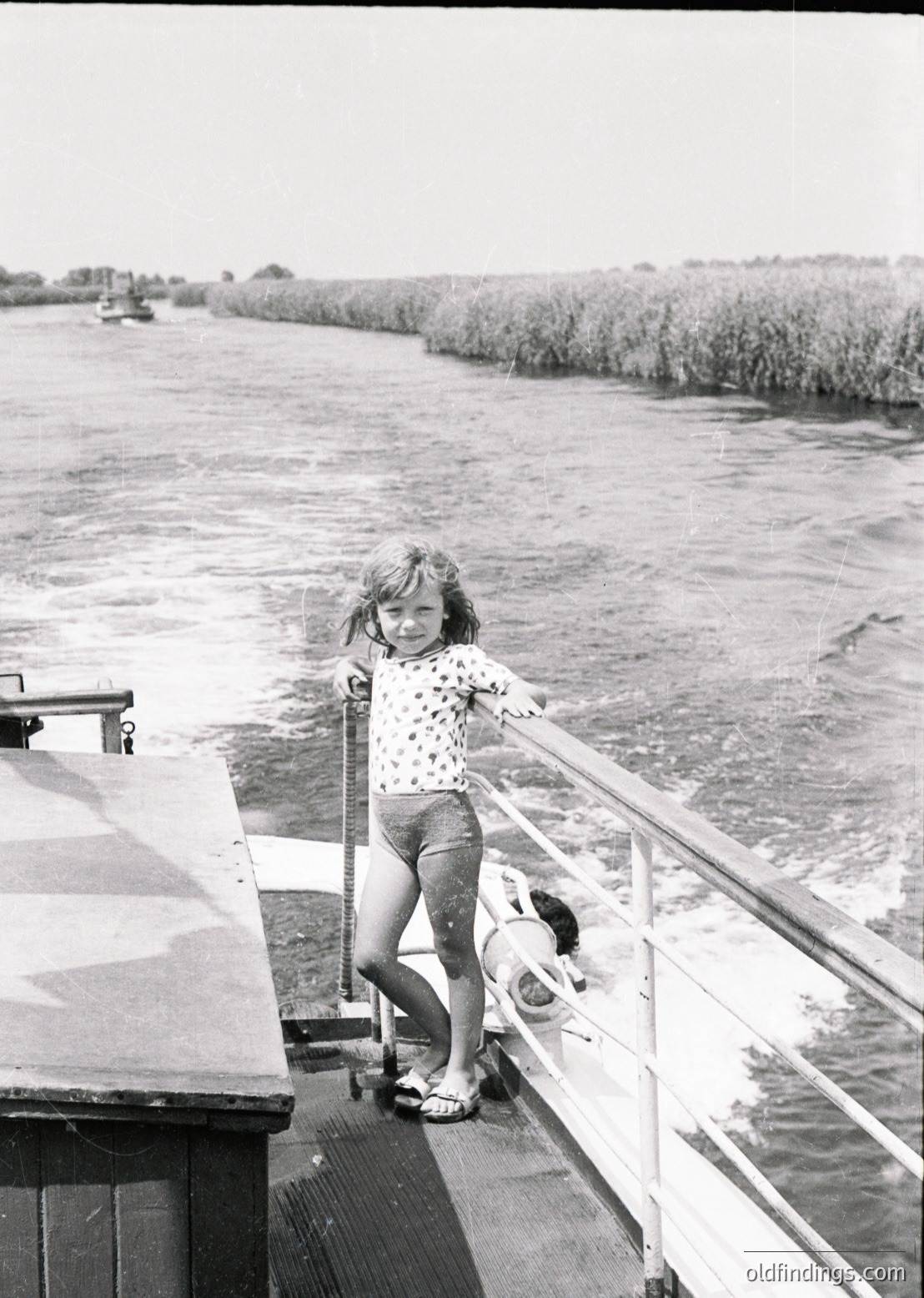 A young girl in a floral dress and sandals stands on a boat railing, smiling at the camera, with reeds and water in the background. Likely a 1960s–1970s river scene, possibly for nostalgic or vintage stock use.