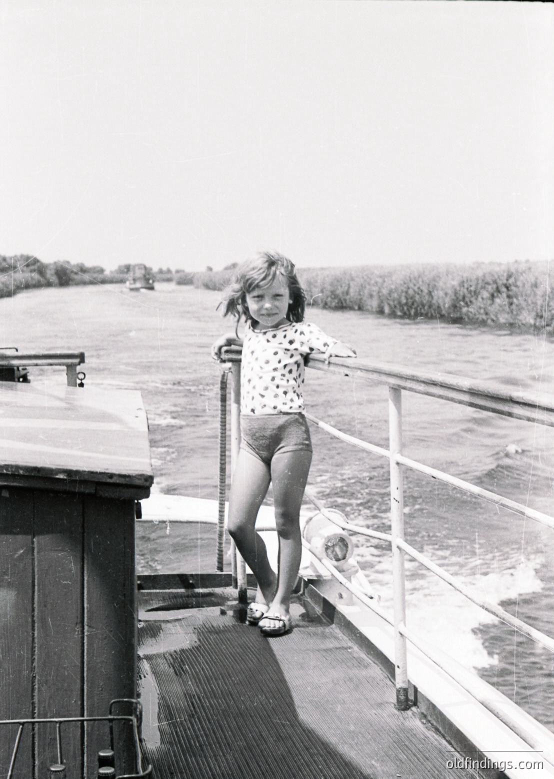 Mid-20th century girl in polka-dot blouse and shorts stands on a ship’s deck railing, gripping metal handrail. Waterway and distant shoreline visible. Classic vintage travel or family photography.