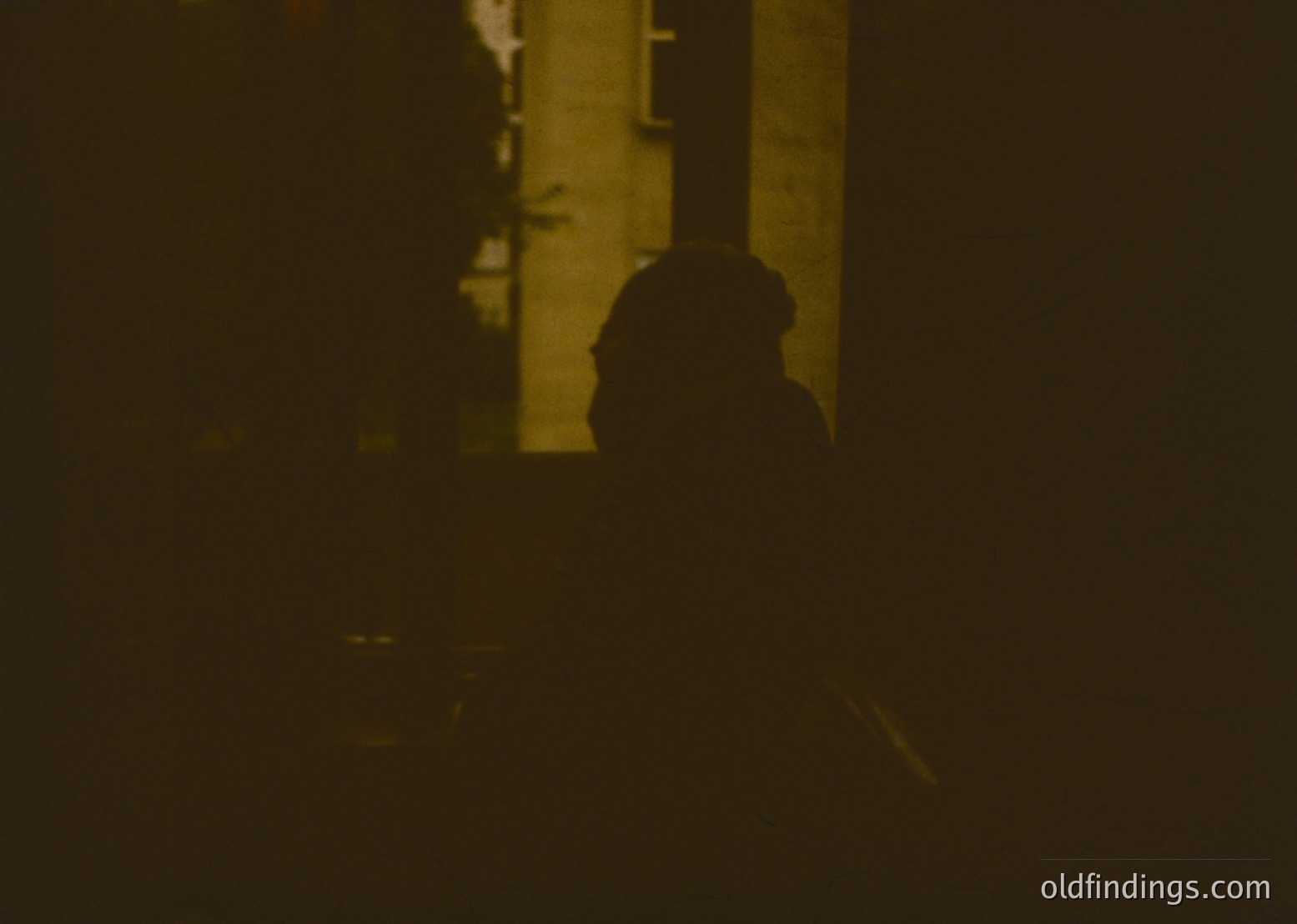Silhouette of a person standing near a dimly lit, narrow alleyway with visible brick walls and a faintly lit doorway. Sepia-toned, low-contrast vintage aesthetic. Likely urban setting, 20th-century street photography.