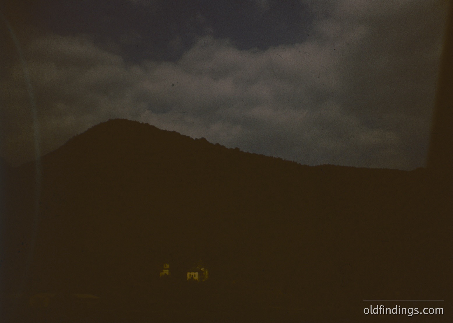 Vintage sepia-toned landscape featuring a lone, isolated house with a sloped roof under dramatic storm clouds. The scene evokes a moody, atmospheric quality, likely from the early-to-mid 20th century.