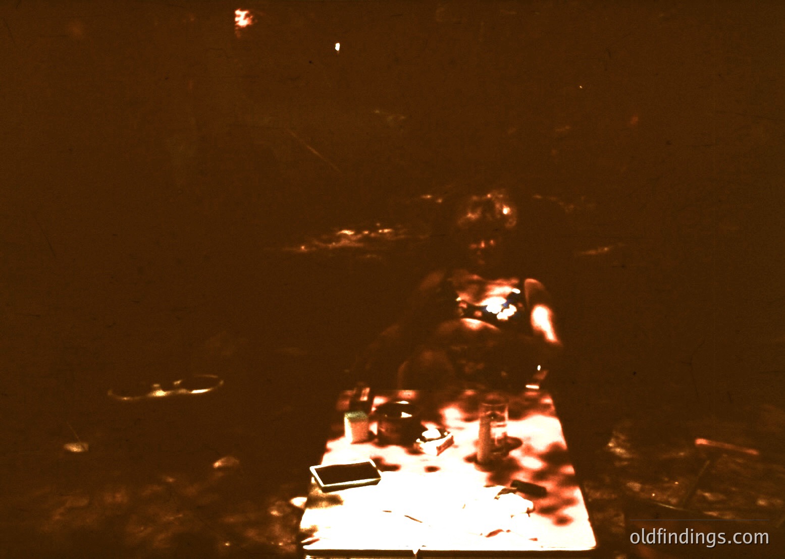 Vintage low-light photo of a dimly lit room with a seated figure at a table. Faded white cloth covers the table, holding a single open book and a glass. Warm, grainy tones suggest analog photography. Likely mid-20th century domestic setting.