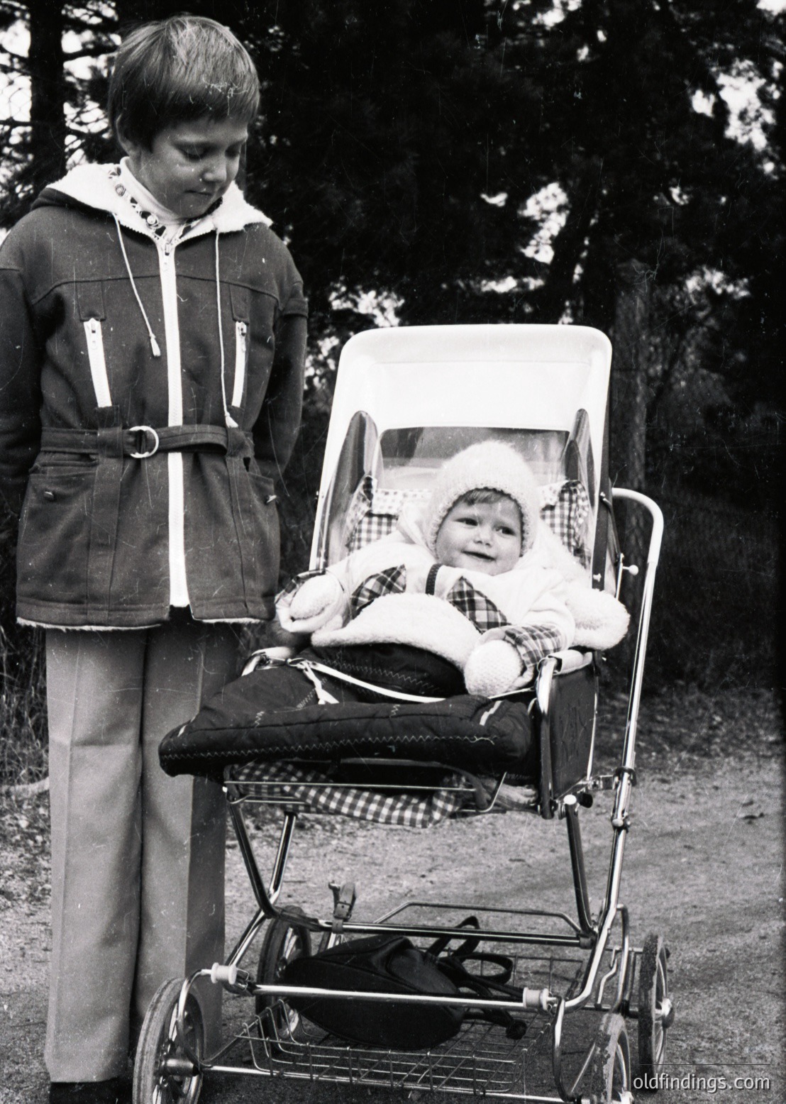 A 1970s-era child pushes a vintage stroller with a baby seated inside, both dressed in winter attire. The older child wears a belted jacket and wide-leg pants, while the baby sports a knitted hat and checkered blanket. Forest foliage frames the scene, suggesting an outdoor park or wooded area.