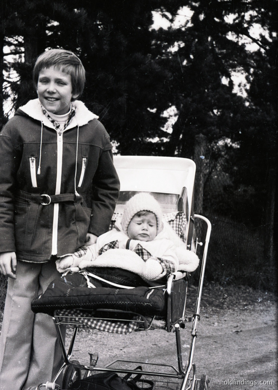 Vintage black-and-white photo of a young boy pushing a baby buggy with an infant dressed in a knitted outfit and hat. The boy wears a zip-up jacket with a belt and wide-leg trousers. Wooded path and dense foliage in the background suggests a park or rural setting, likely mid-20th century.