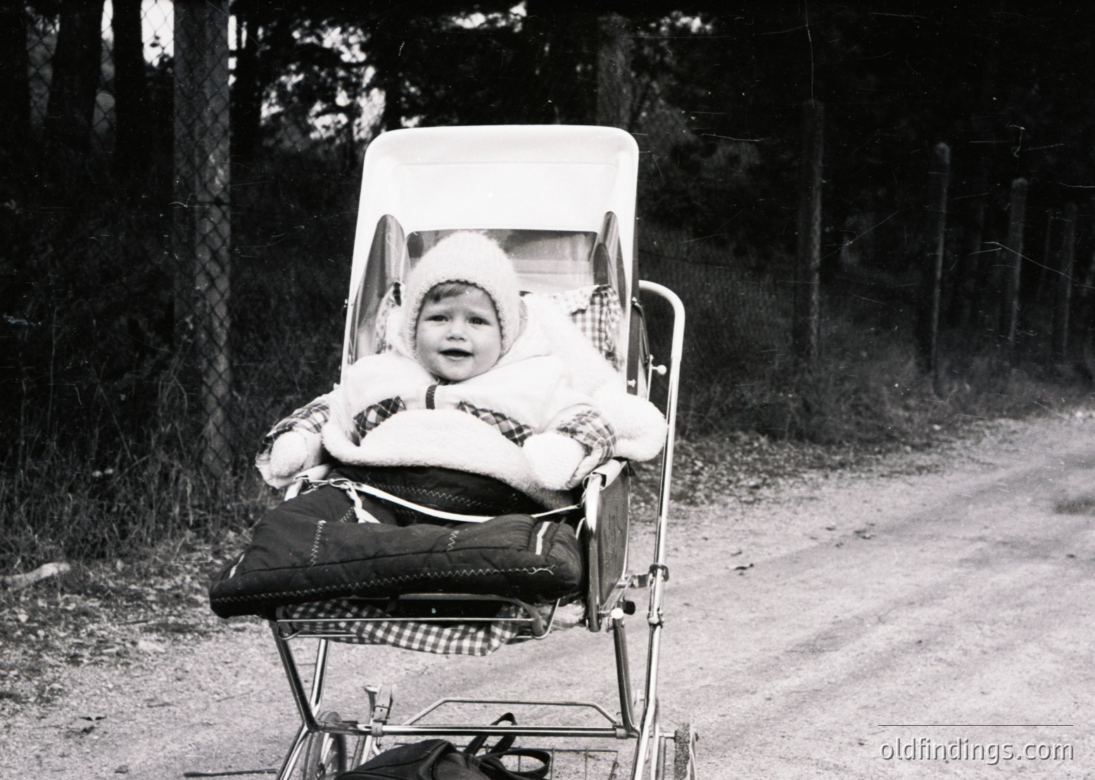 Vintage black-and-white photo of a child in a vintage baby carriage on a rural road, bundled in a knitted hat and fur-lined coat. Metal frame with woven seat detail. Mid-20th century rural lifestyle.