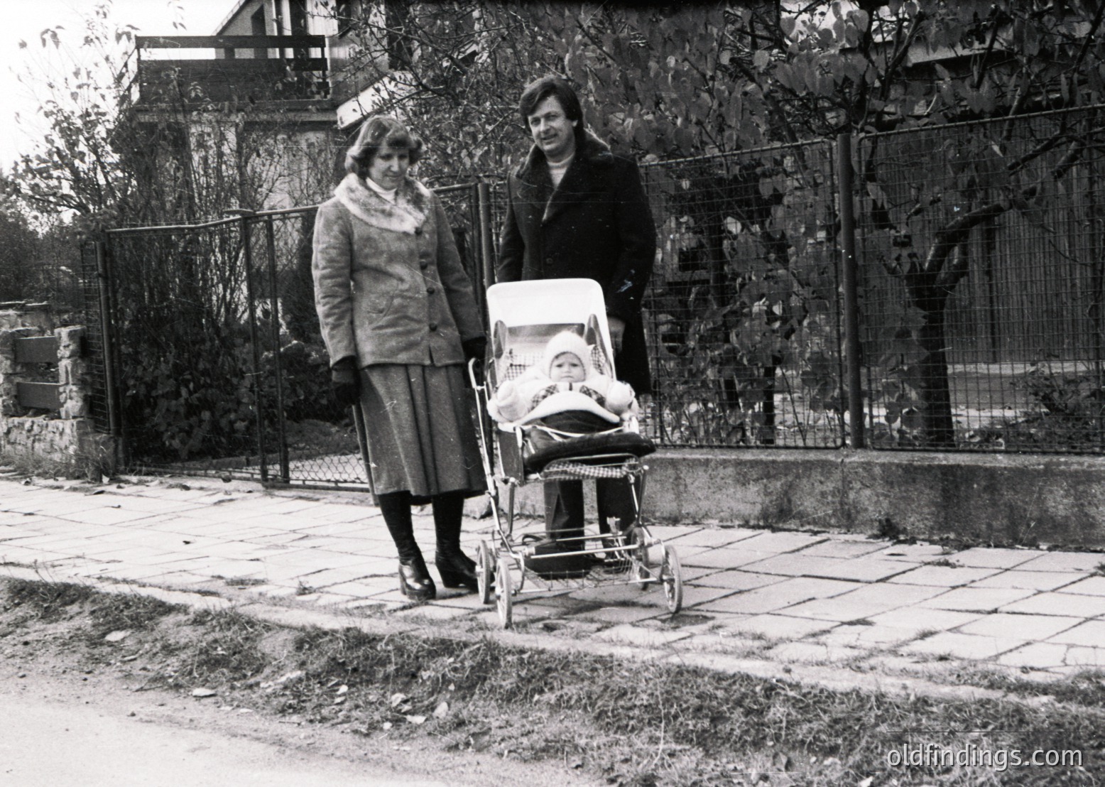 Two women in 1960s-era attire pose outdoors beside a classic metal-framed baby stroller. The woman on the left wears a fur collar coat and skirt; the other, a hat and coat, holds the stroller’s handle. A child sleeps inside. Residential fence and paved sidewalk suggest a suburban neighborhood.