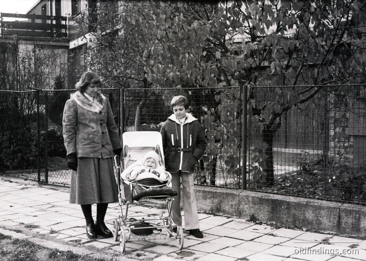 A mid-20th-century black-and-white snapshot captures a woman in a knee-length coat and scarf pushing a vintage pram, accompanied by a young boy in a puffy jacket and wide-leg trousers. The scene appears outdoors, likely in a residential area with a chain-link fence and leafy trees in autumn. The composition evokes 1960s–1970s family life.