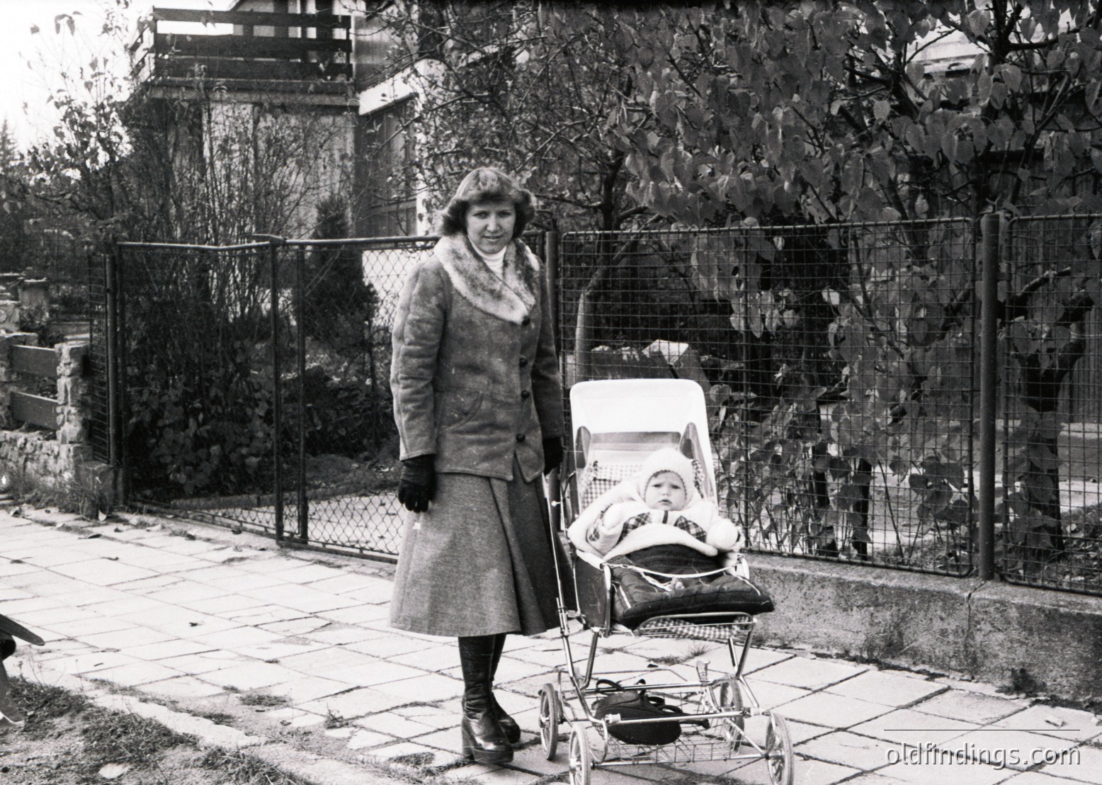 A woman in 1960s-era attire—coat, gloves, and hat—pushes a classic metal-framed pram with a sleeping infant. Residential street with chain-link fence and autumn foliage in background.