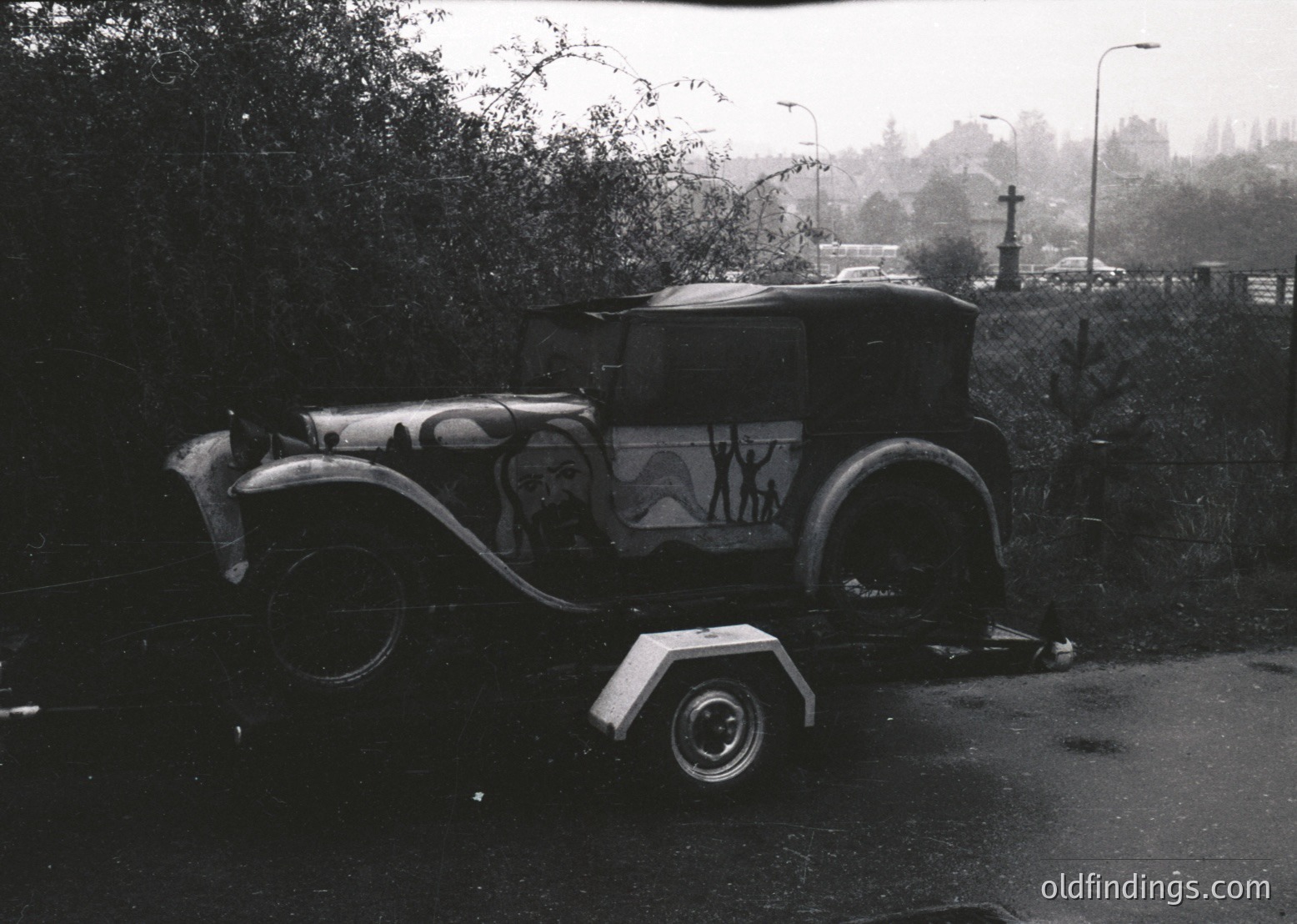 Vintage flatbed trailer towing a 1930s-era sedan with decorative mural depicting human figures, parked on a wet roadside. Surrounding greenery and a cemetery with gravestones in background. Classic mid-century American rural scene.