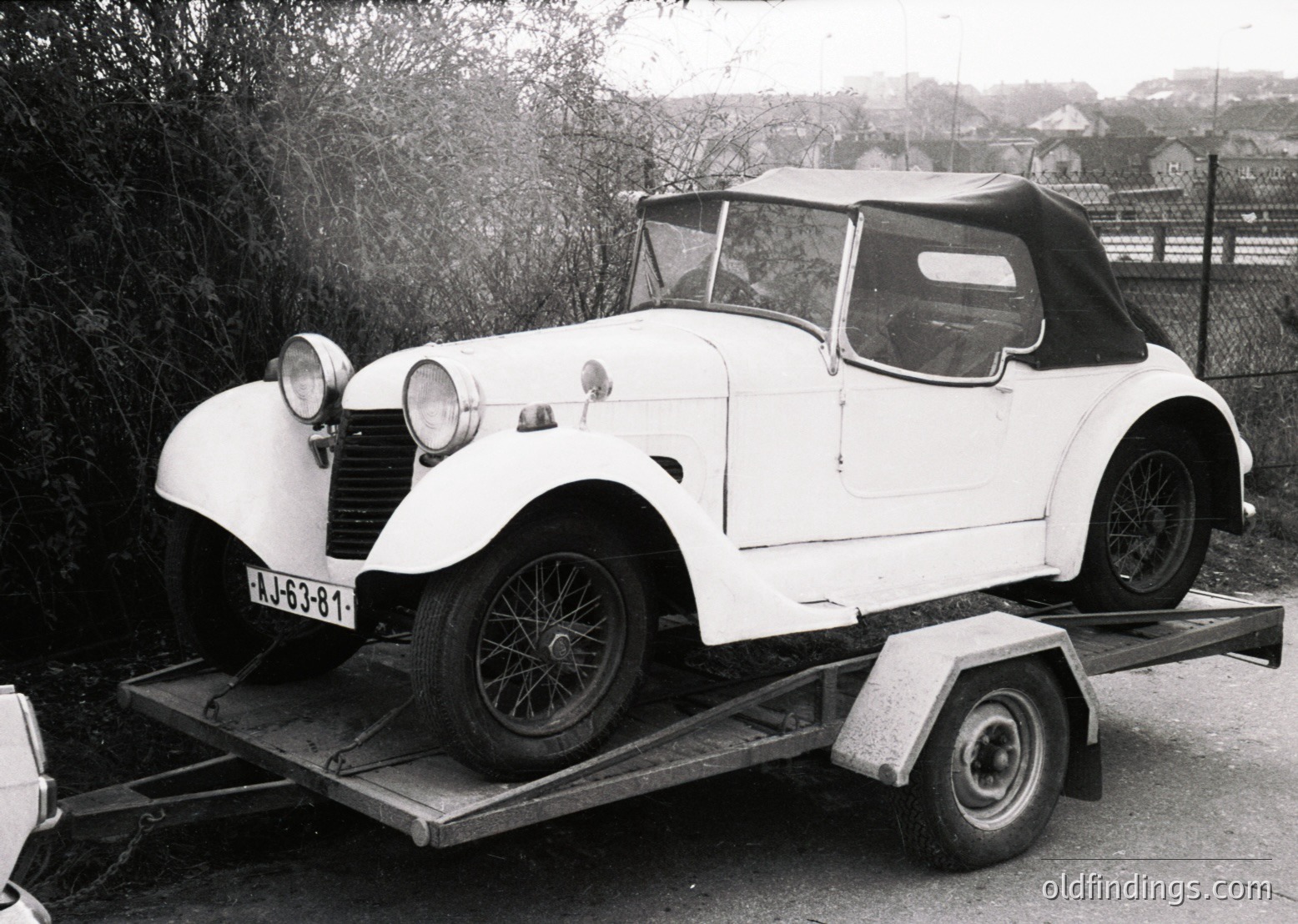 Classic white convertible roadster on trailer, likely a 1930s–1940s model, featuring rounded fenders, dual headlamps, and a vinyl top. Rustic rural setting with sparse trees and a fence in background. License plate "A-53-31" suggests Eastern European origin.