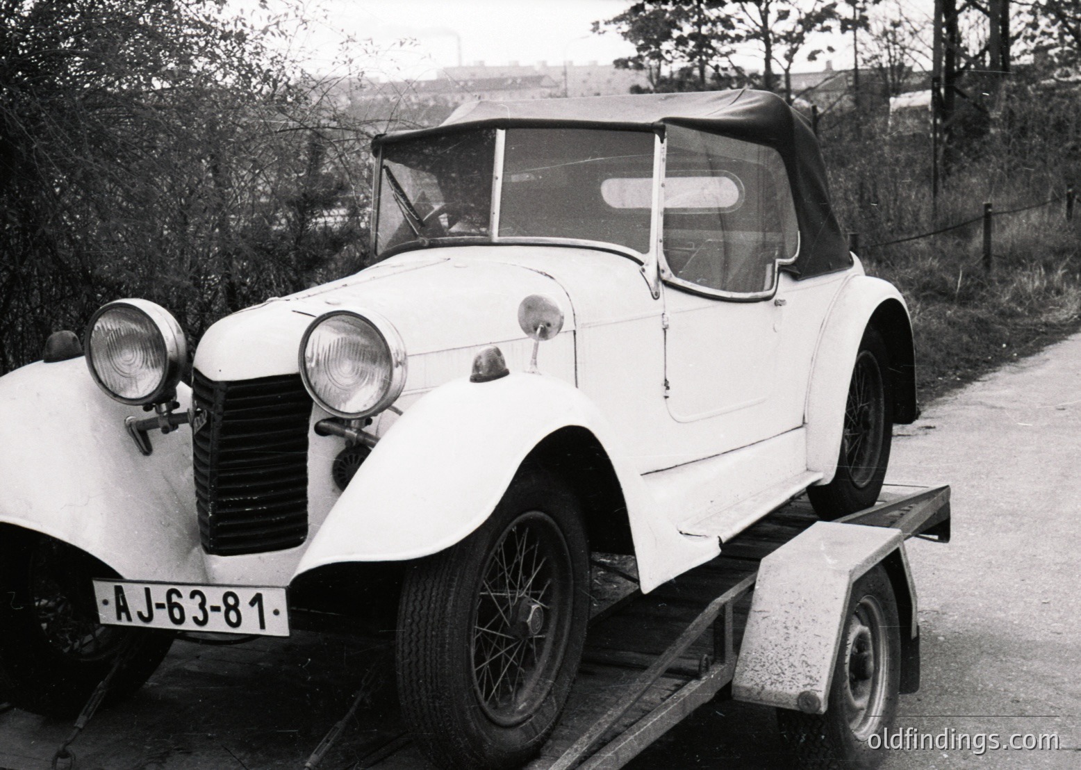 Vintage white convertible car (likely 1930s-1940s) on a trailer, featuring rounded fenders, dual headlamps, and a soft-top roof. License plate "AJ-63-81" suggests Eastern European origin. Rustic roadside setting with sparse trees and distant buildings.