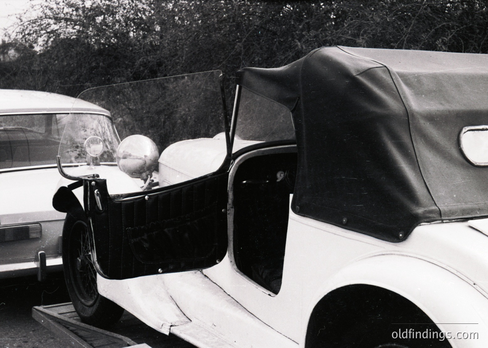 Vintage convertible with classic rounded fenders, whitewall tires, and a rolled-down leather roof, parked beside a utilitarian sedan. Distinctive round headlight and chrome detailing suggest a mid-20th-century design, likely 1950s–1960s. Open door reveals interior trim.