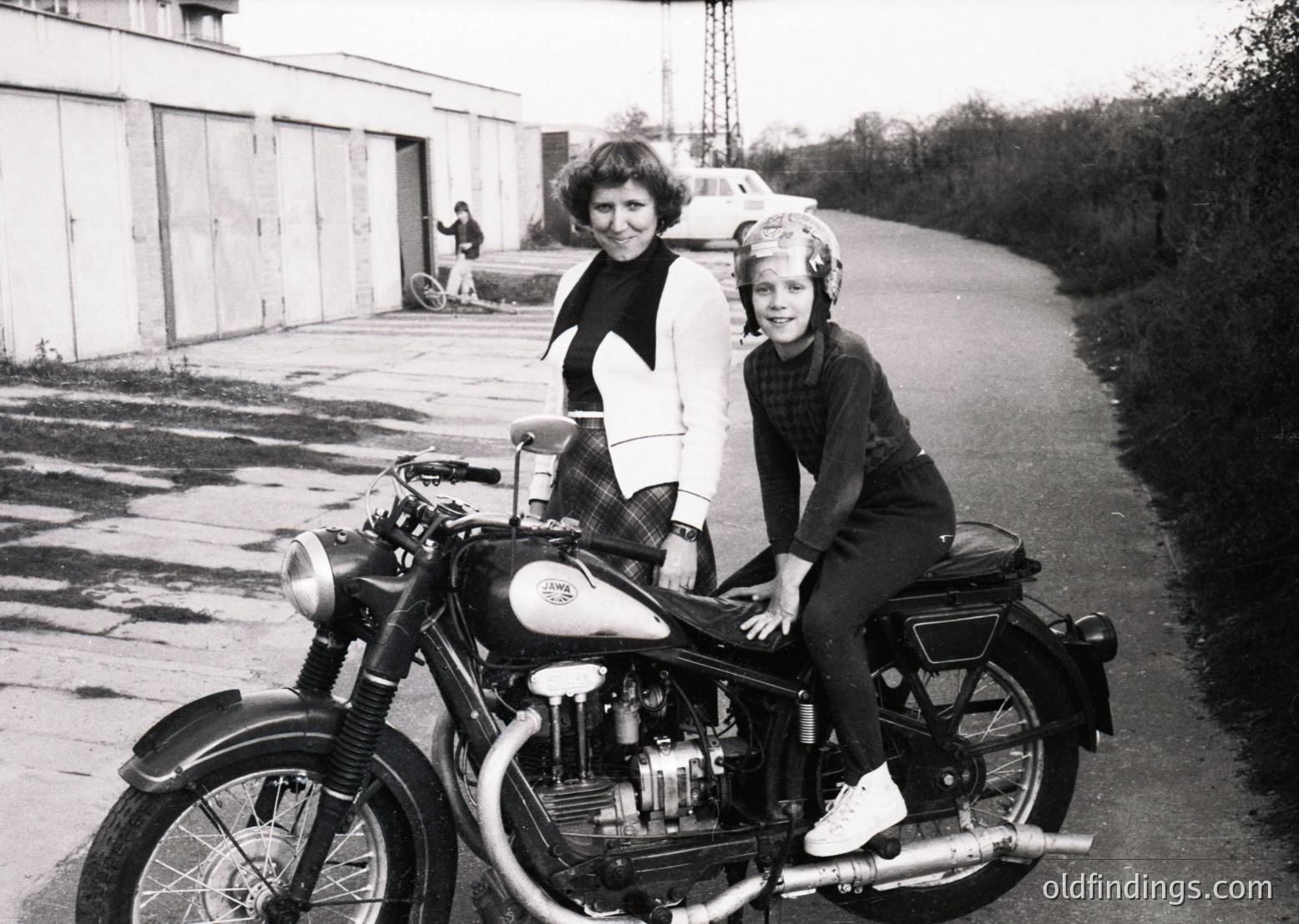 Vintage black-and-white photo of a woman and child seated on a classic motorcycle, likely a BSA or similar British model, in an industrial setting. The woman wears a patterned sweater and skirt; the child, a striped sweater and helmet. Background shows a garage door and parked cars, suggesting mid-20th century (1950s–1960s) urban or suburban Europe/UK.