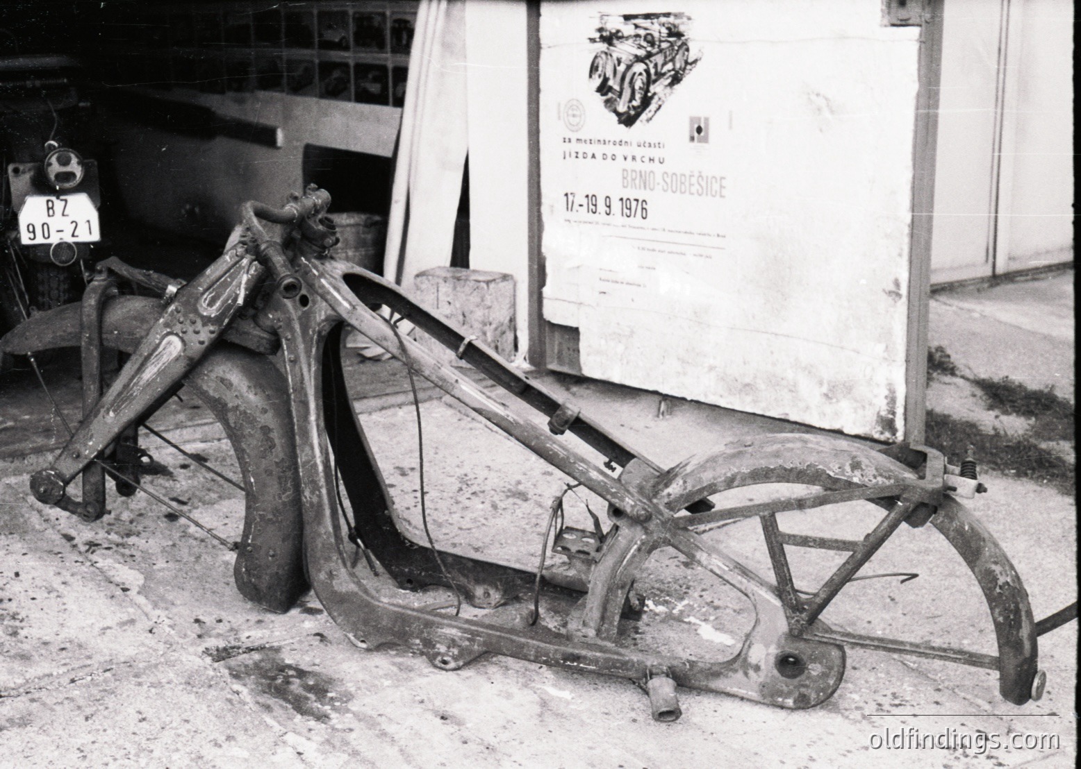 Vintage motorcycle frame in disassembled state, likely for repair or restoration, with visible welding seams and rust. Poster in background advertises a 1976 event in Czech (likely a motorcycle rally or exhibition). Industrial workshop setting with partial license plate () visible.