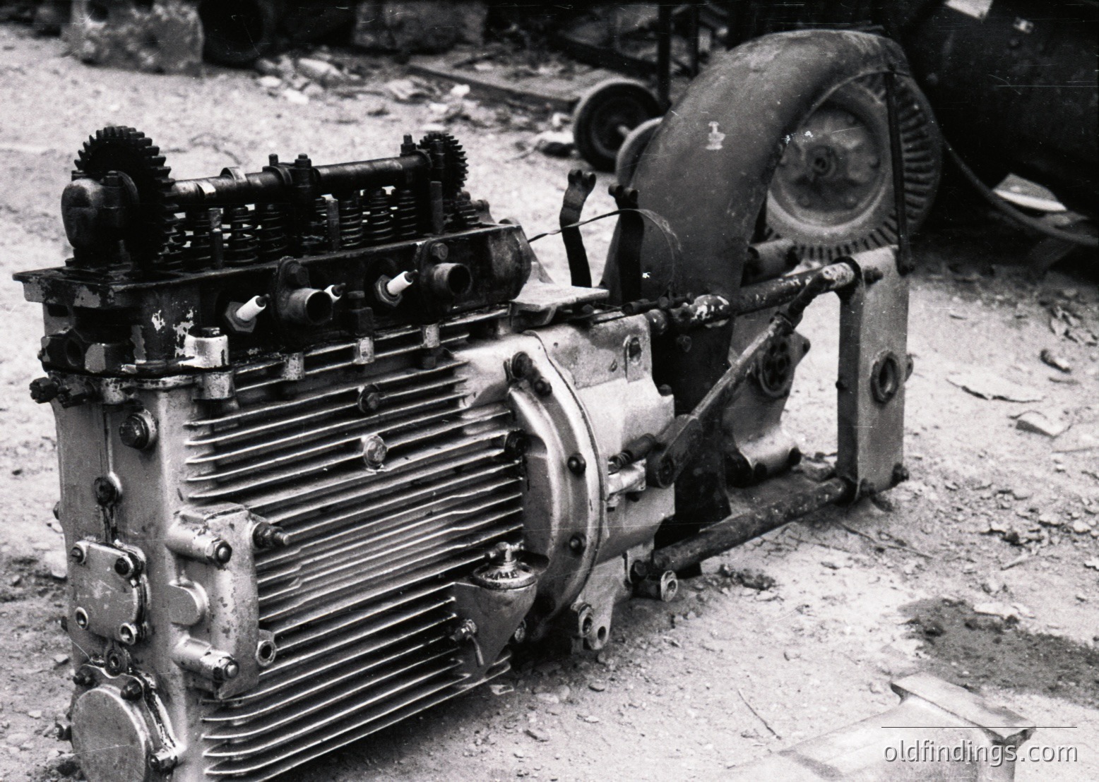 Vintage industrial engine with exposed cylinder fins, gearing, and flywheel, likely from mid-20th century machinery. Rust and dirt suggest outdoor or workshop use. Potential automotive or stationary power application.