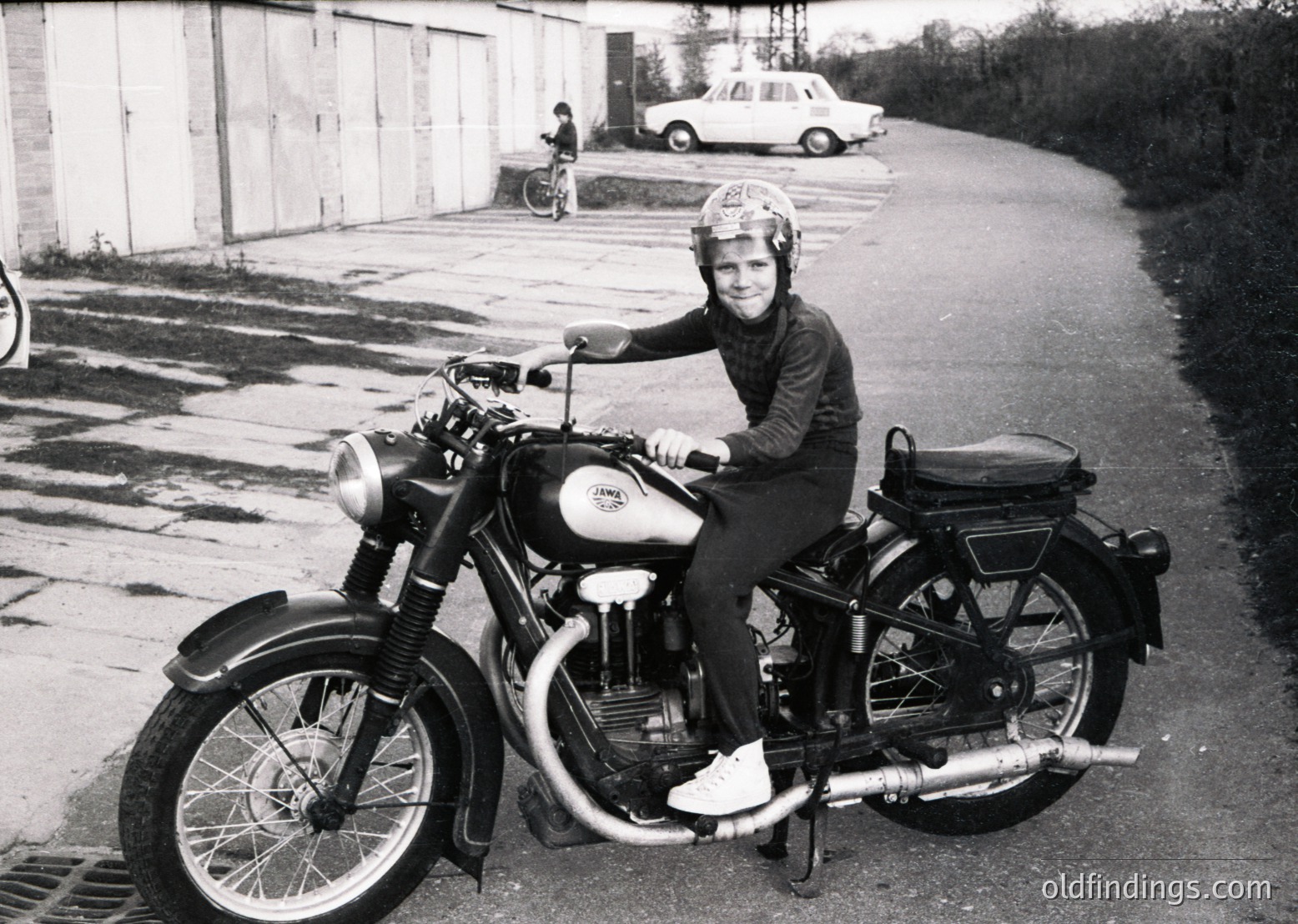 Mid-century vintage motorcycle rider in protective gear, likely 1950s–1960s. Classic CWK or CWK-style bike with chrome details and sidecar seat. Industrial background suggests urban or suburban setting. Black-and-white photo captures mid-20th-century transportation culture.