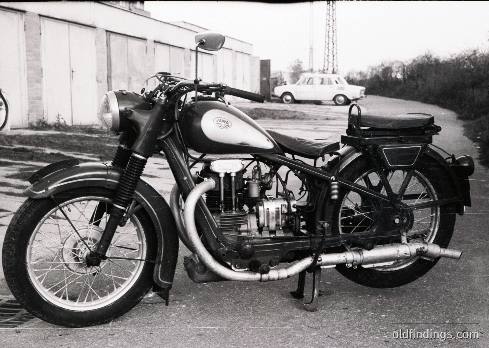 Classic Jawa motorcycle, likely a **Jawa 350cc** model, photographed in mid-20th century (1950s–1960s). Prominent sidecar-mounted engine with exposed valves, dual headlamps, and spoked wheels. Industrial setting with parked vintage car in background.