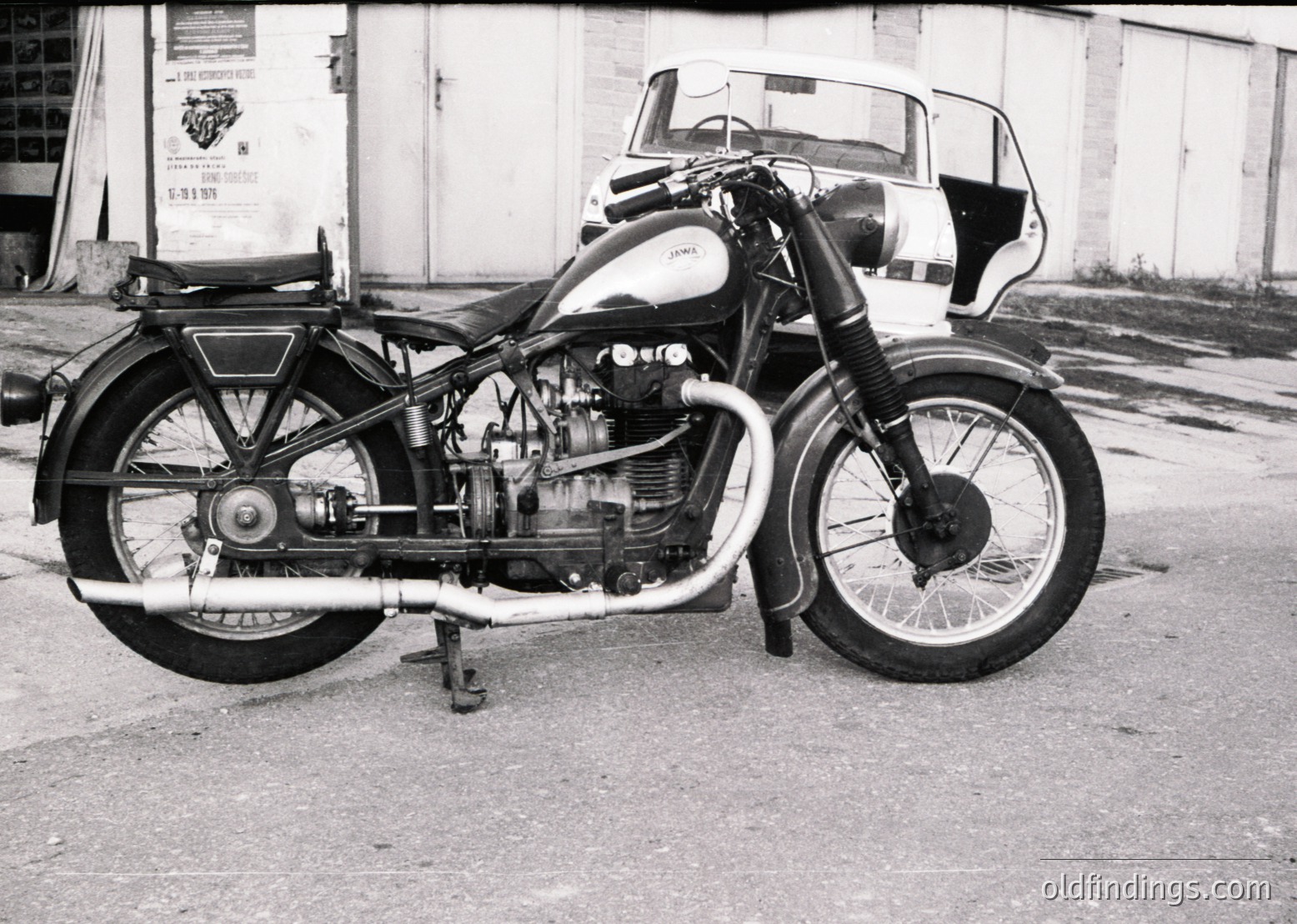 Classic **AJS Model 20/25 sidecar motorcycle** (1950s–1960s) with exposed V-twin engine, pressed-steel fenders, and spoked wheels. Sidecar features a streamlined passenger compartment. Industrial setting suggests vintage showroom or garage.