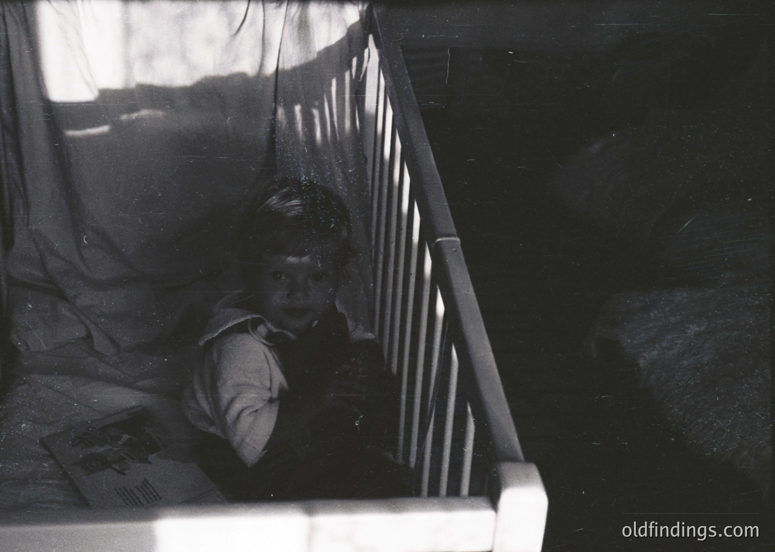 Black-and-white candid of a child peering through a crib’s vertical bars, clutching a booklet with a visible illustration. Soft indoor lighting highlights the child’s focused expression. Likely mid-20th century domestic setting, evoking nostalgia for childhood reading habits.