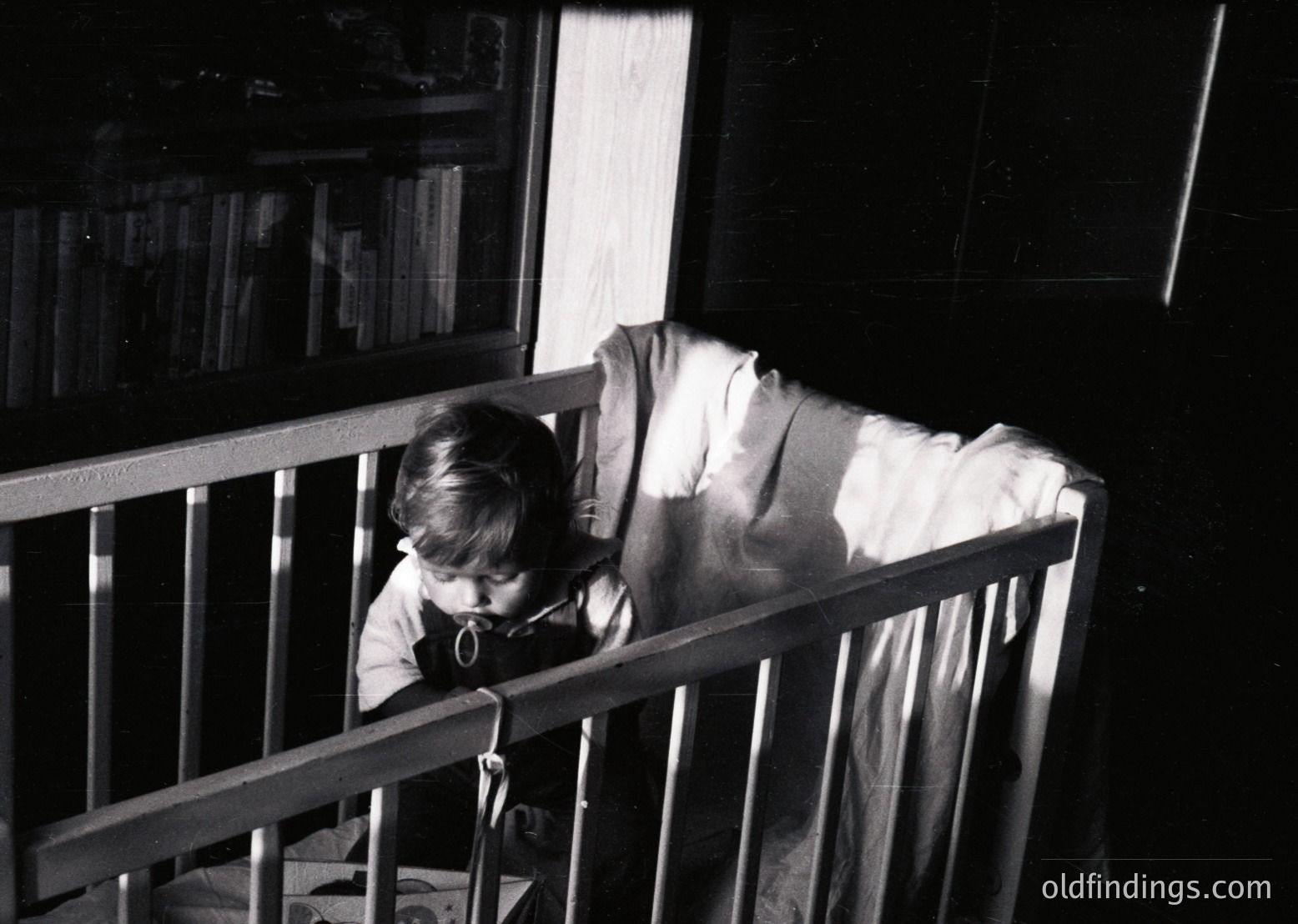 A young child sits in a wooden crib, leaning forward with hands on the bars, gazing intently. Bookshelves filled with books line the dimly lit room’s wall, illuminated by a single window. Mid-20th century domestic interior, likely 1950s–1960s.