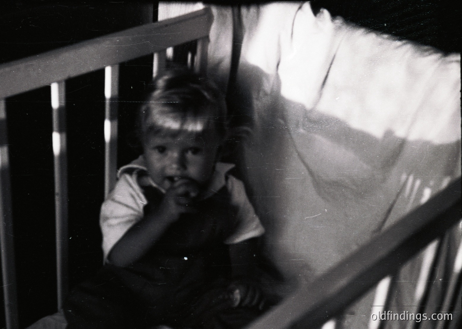 Young child in vintage striped shirt peering through metal balcony railing, mid-20th century. Soft focus and grainy texture suggest early film photography. Indoor/outdoor transition with blurred background light.