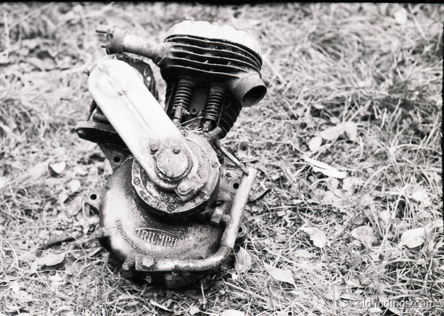 Vintage Triumph motorcycle engine, mid-20th century, resting on grassy ground. Rust and wear highlight its aged condition, with visible "Triumph" branding on the cylinder. Classic British motorcycle heritage.