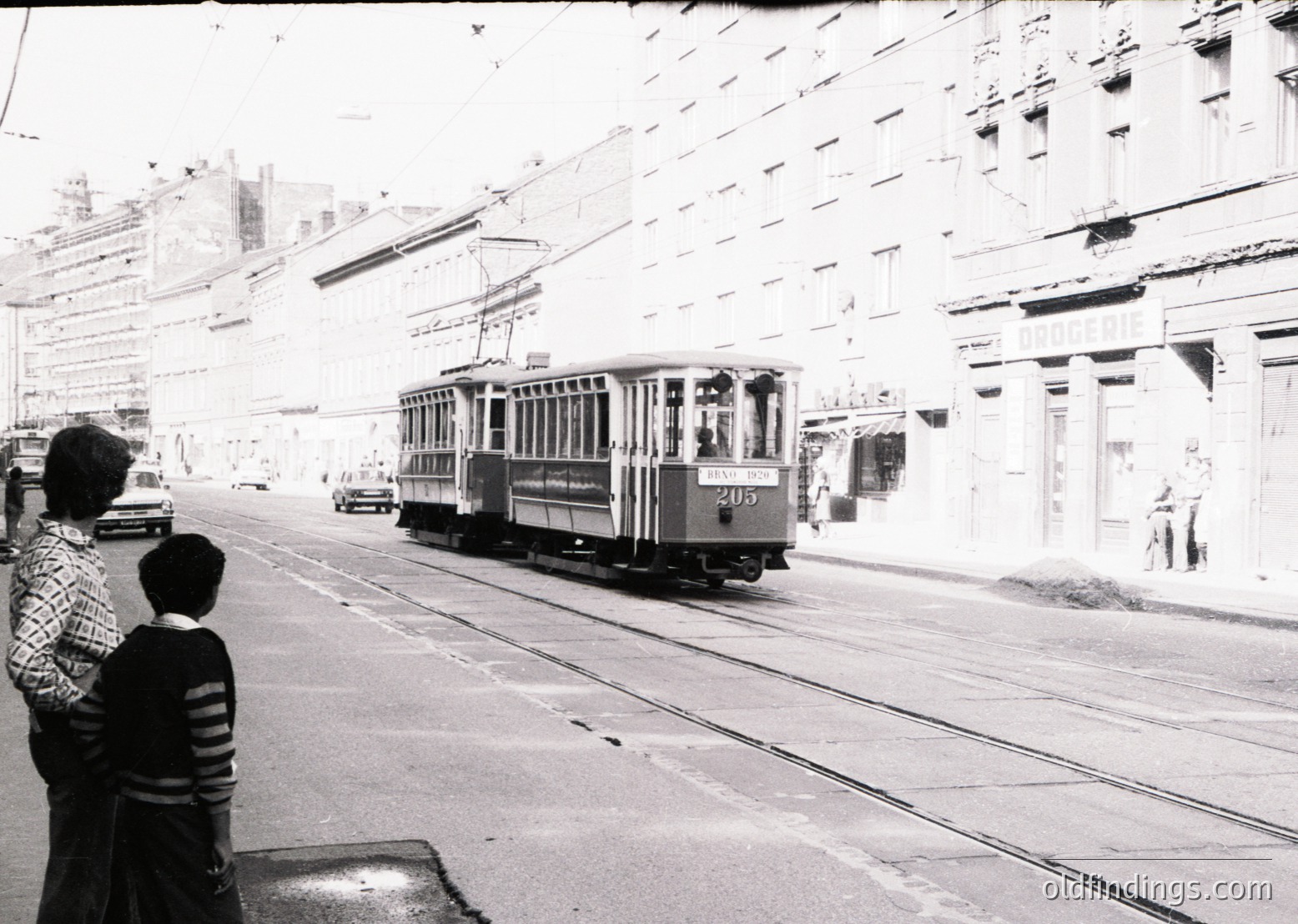 Vintage streetcar () navigating urban tracks in mid-20th century cityscape. Pedestrians observe from sidewalk beside mid-rise buildings with balconies. Overhead wires and classic tram design suggest European transit system, likely 1950s–1960s.