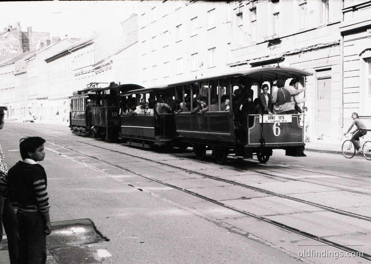 Vintage tram with open-air seating, likely a **1970s European urban transit system**, navigating a steep incline. Pedestrians and cyclists share the street with mid-rise residential buildings. Classic streetcar design reflects mid-century public transport.