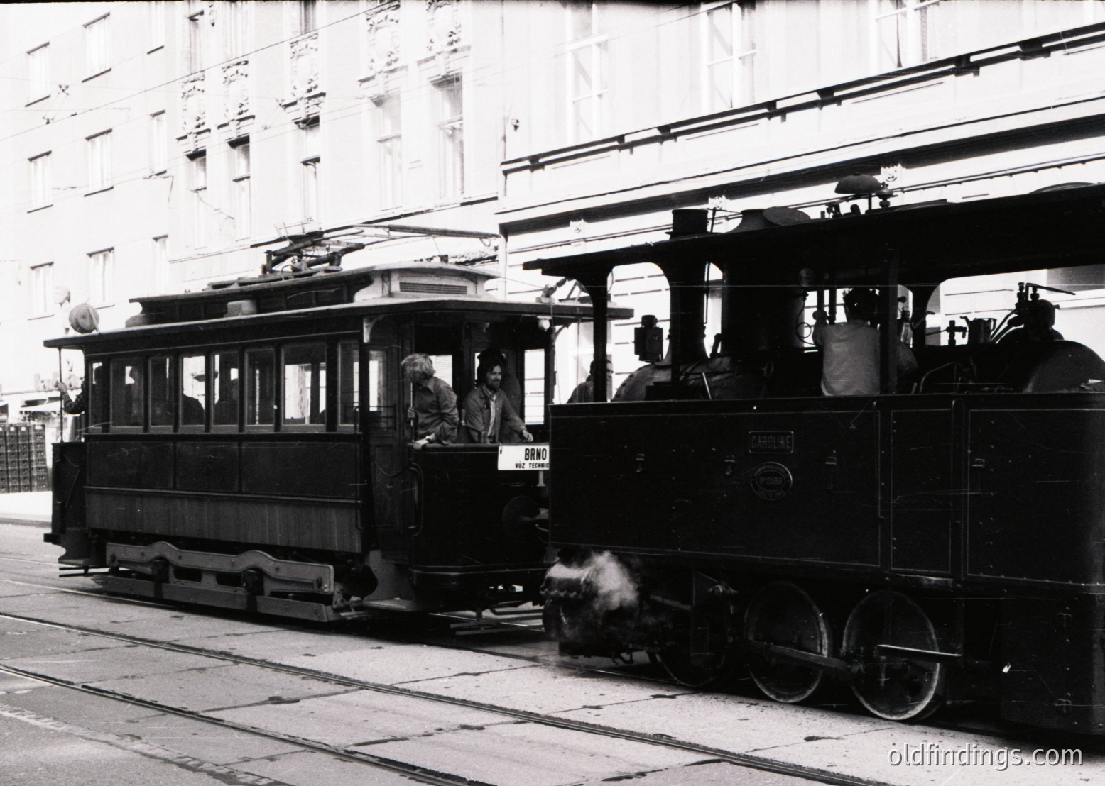 Vintage streetcar with open platform and wooden construction, likely a 1920s–1940s urban transit vehicle. Steam-powered locomotive in background suggests mixed rail/tram infrastructure. Architectural details hint at European cityscape, possibly Eastern Bloc era.