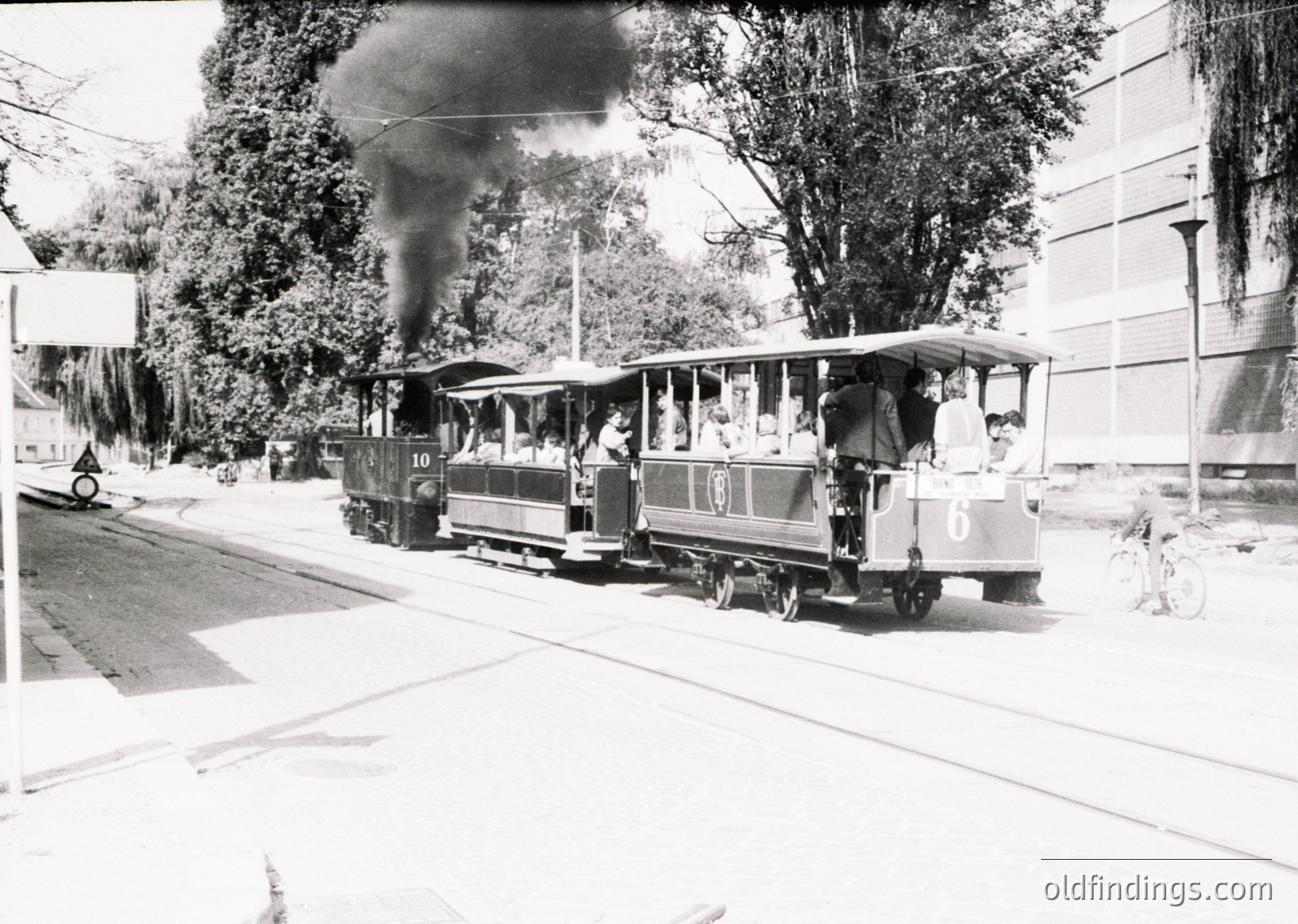 Vintage steam-powered tram with numbered cars (10 & 6) traveling on urban tracks, emitting visible smoke. Passengers stand on open platforms; trees and a building flank the street. Likely mid-20th century European city.