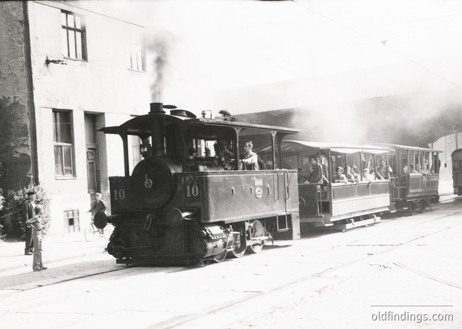 Vintage steam locomotive pulling open-air passenger cars, likely a tourist train, in early 20th-century Europe. Smoke billows from the stack, and passengers stand or sit in open carriages. Nearby, a man in uniform directs traffic at a station platform with classic brick buildings.