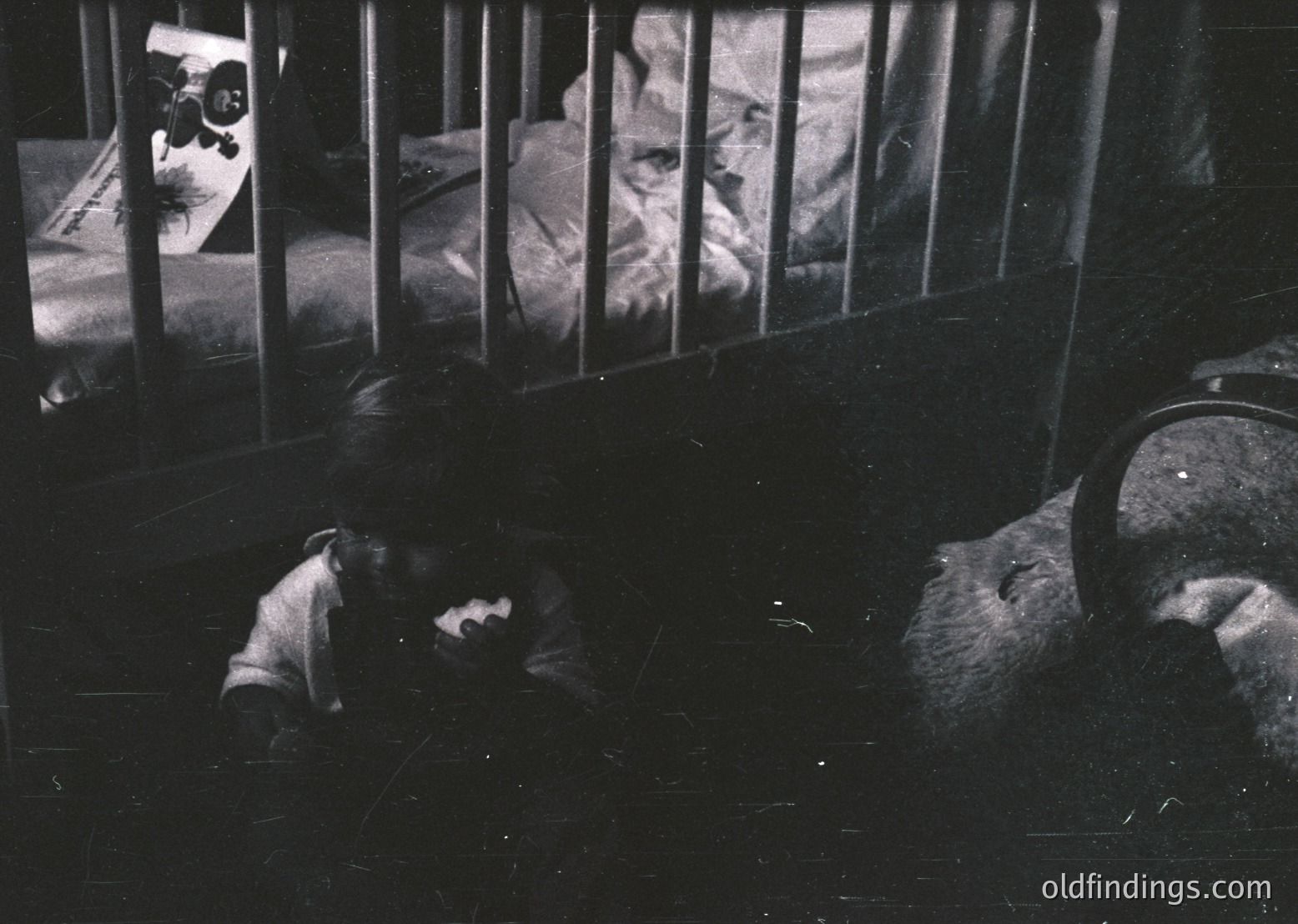 Black-and-white prison cell interior with barred window revealing another inmate seated on a bunk. Visible are a metal bed frame, a small window with vertical bars, and a reflection of a guard or officer in the glass. Mid-20th century institutional design.