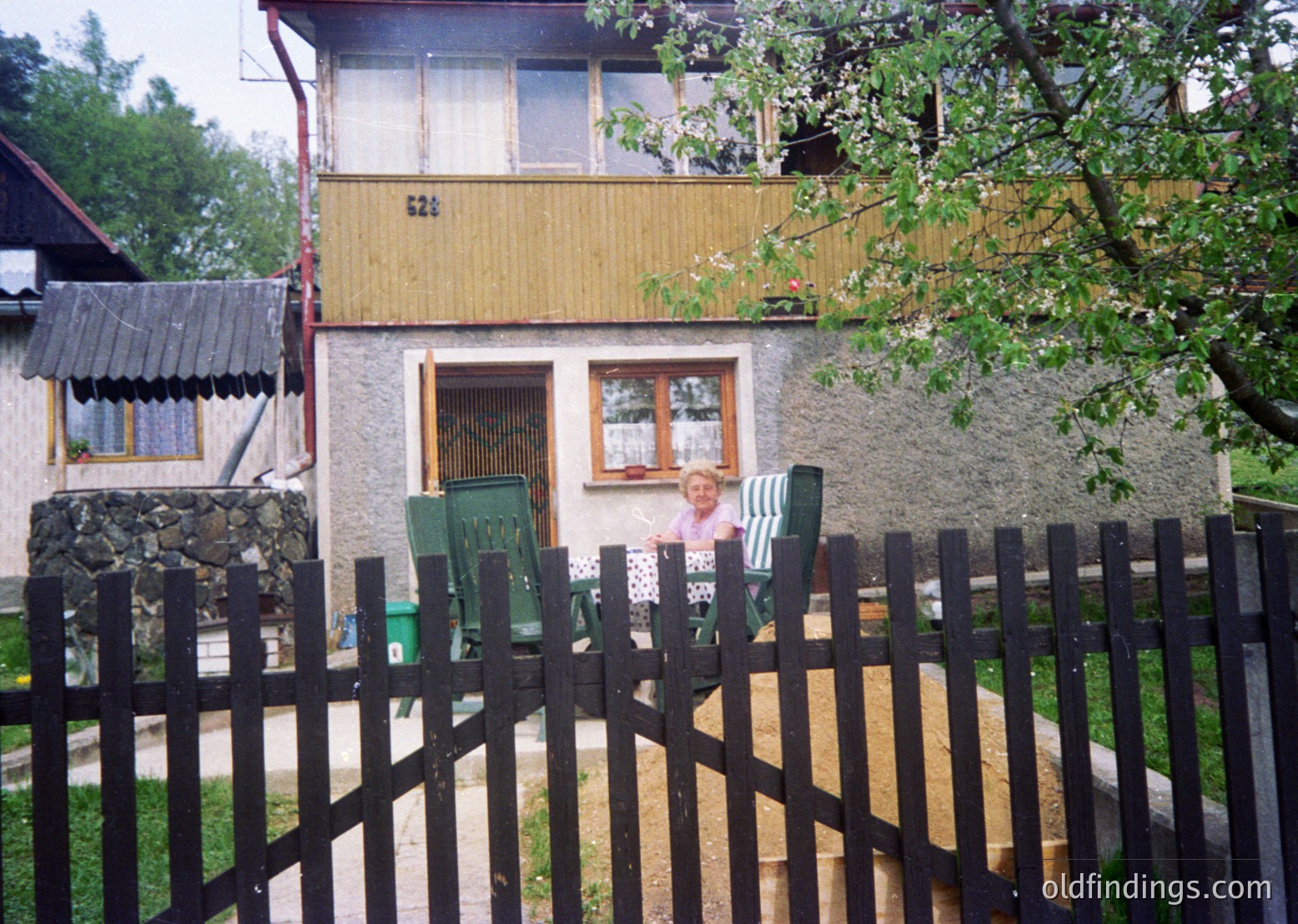 Two children pose in front of a mid-20th-century residential house with a yellow upper floor and grey lower walls. Green plastic chairs and a stone foundation detail the front yard, framed by a black metal fence. Lush greenery and a rustic shed add to the rural setting.