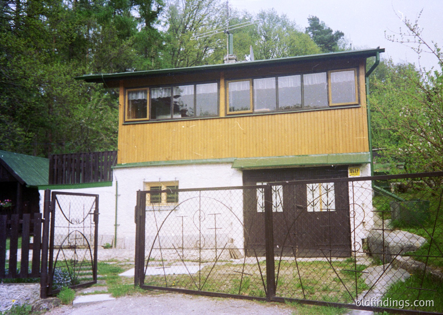 Mid-century modern wooden house with yellow upper floor and white lower level, featuring large windows and a gabled roof. Wrought-iron gate with arched design frames the entrance. Surrounded by dense greenery, suggesting a rural or suburban setting.
