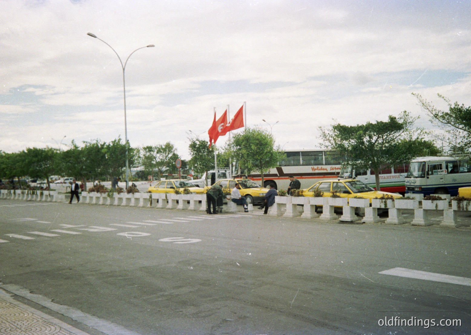 Urban scene featuring a busy roundabout with yellow taxis, a prominent Turkish flag, and Soviet-era architecture in background. Likely Istanbul, 1980s-1990s.