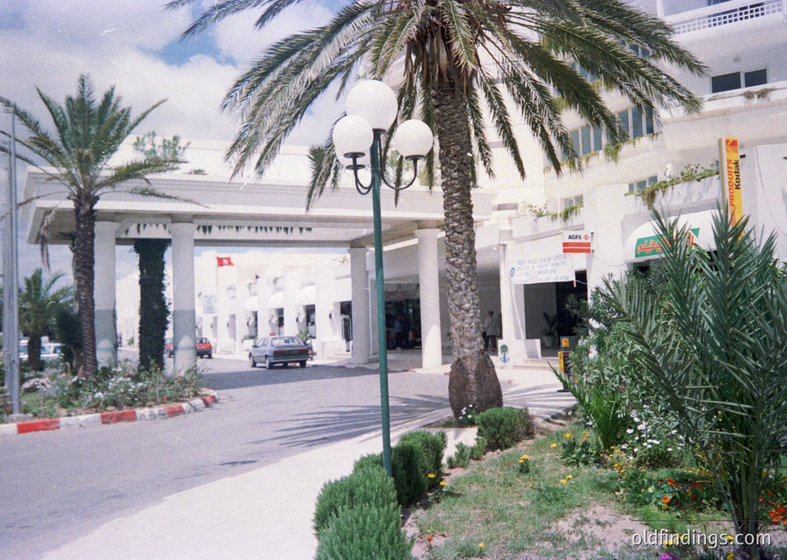Neatly landscaped Mediterranean-style entrance with tall palm trees, ornate street lamps, and a grand archway. Mid-20th century resort architecture featuring whitewashed walls, wrought iron accents, and vibrant signage ( )